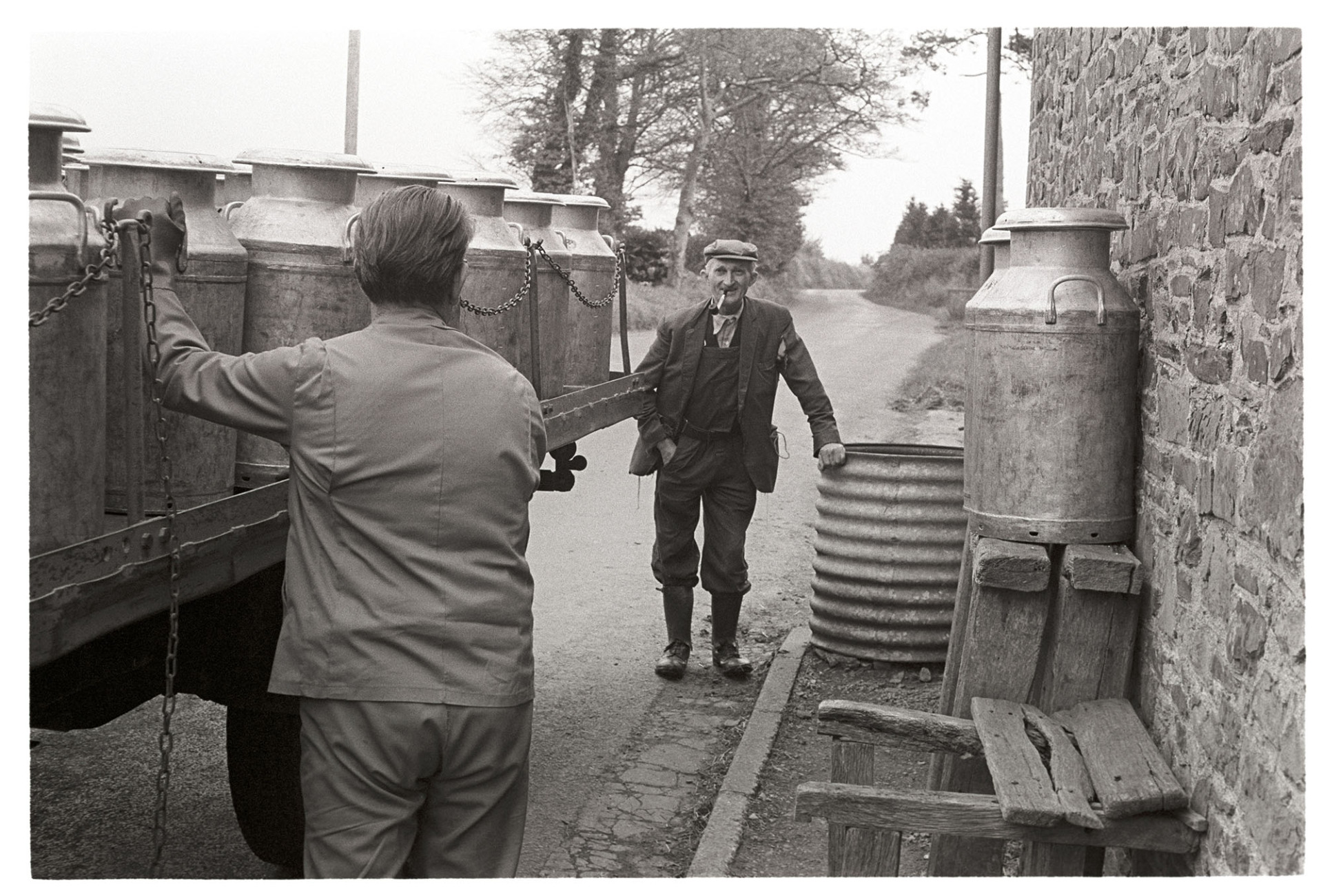 Milk lorry arriving to pick up milk churns, farmer chatting to driver. 
[Gordon Sanders smoking a pipe and talking to a milk lorry driver who has arrived to pick up milk churns at Reynards Park, Ashreigney.]