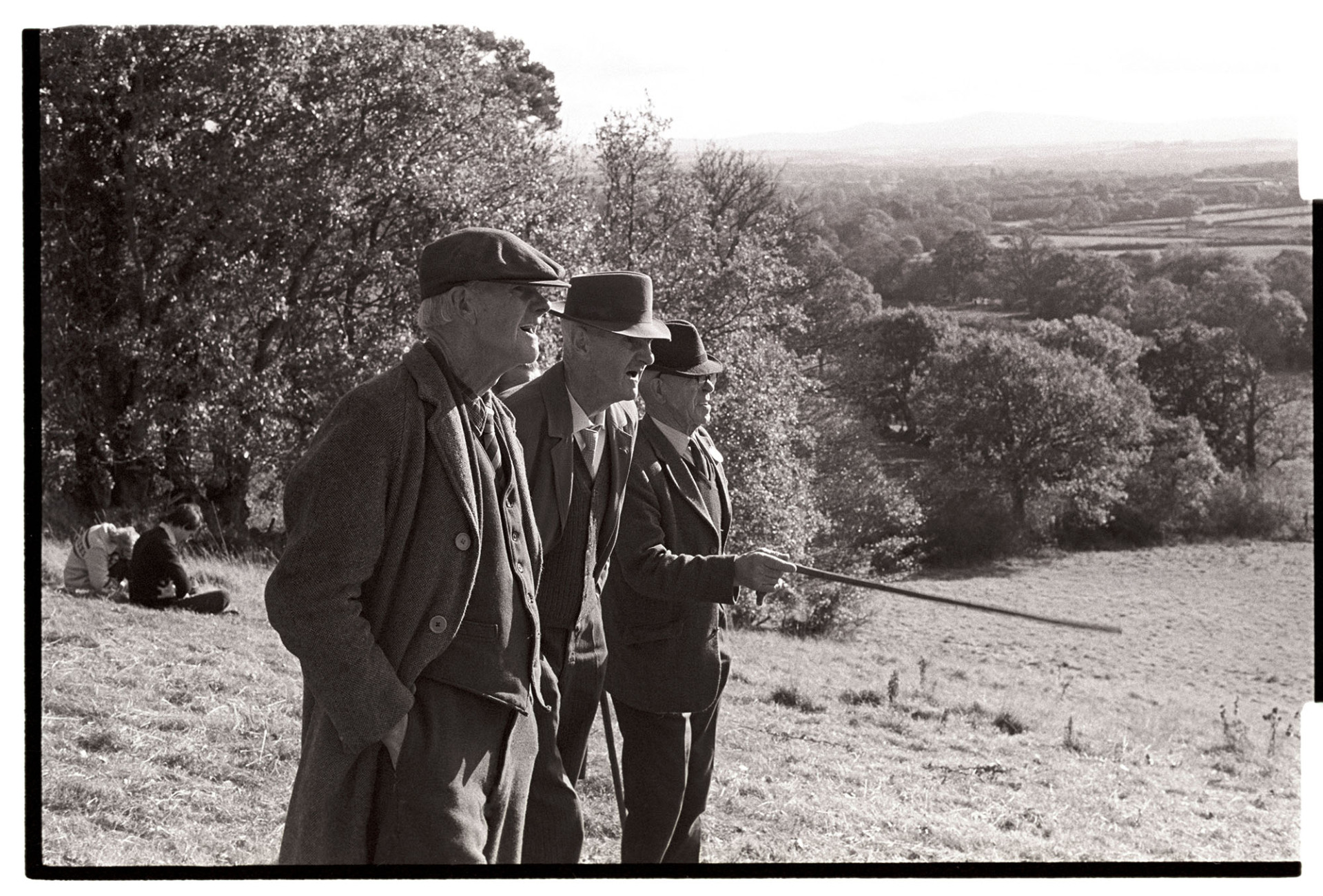 Hunt followers, three men. 
[Three men stood in a field on a hillside watching a hunt, near Hatherleigh. One of the men is pointing with his walking stick. Two other people are sat by some trees in the background.]