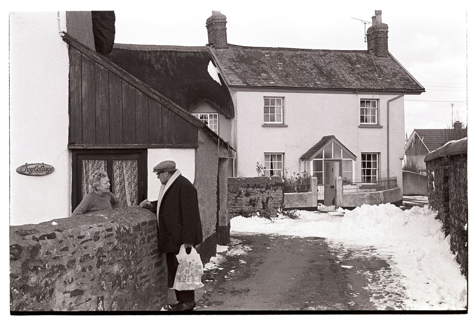 Snow, couple chatting over wall, house with unusual roof. 
[A man and woman talking over a wall at Joy Cottage in Dolton. In the background a snow covered street and cottage with a slate roof is visible. The roof has an overhang at one end.]