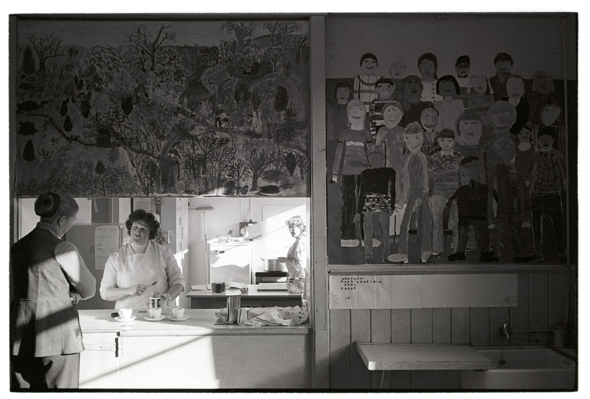 Teacher and school dinner lady chatting at counter of canteen with large paintings, murals. 
[A teacher and dinner lady talking at the serving hatch of the school canteen at Burrington Primary School. Children's schoolwork is displayed by the serving hatch.]