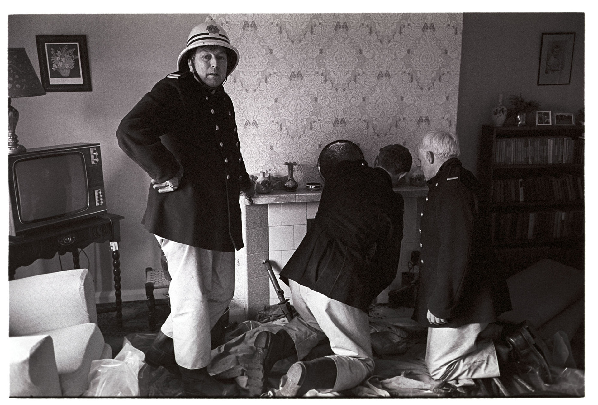 Firemen at chimney fire, clearing out mess in sitting room fireplace. 
[Firemen from Hatherleigh Fire Brigade attending a chimney fire at a house in West Lane, Dolton. They are clearing the fireplace in the sitting room of the house. A chair, television and patterned wallpaper are visible in the room.]