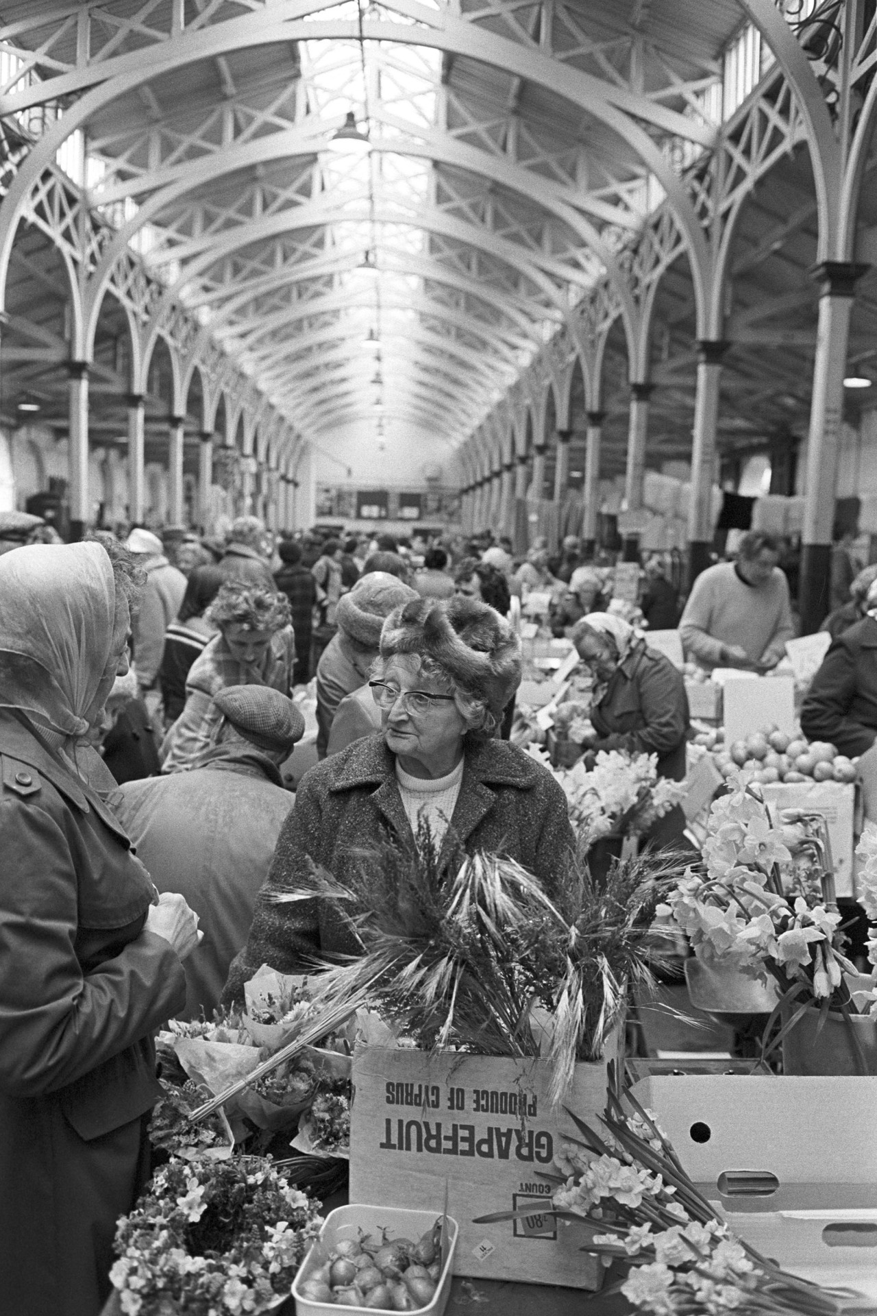 Pannier market, woman vegetable and flower stall. 
[Two women talking at a stall with flowers and onions at Barnstaple Pannier Market. Other market traders and shoppers are visible in the background.]