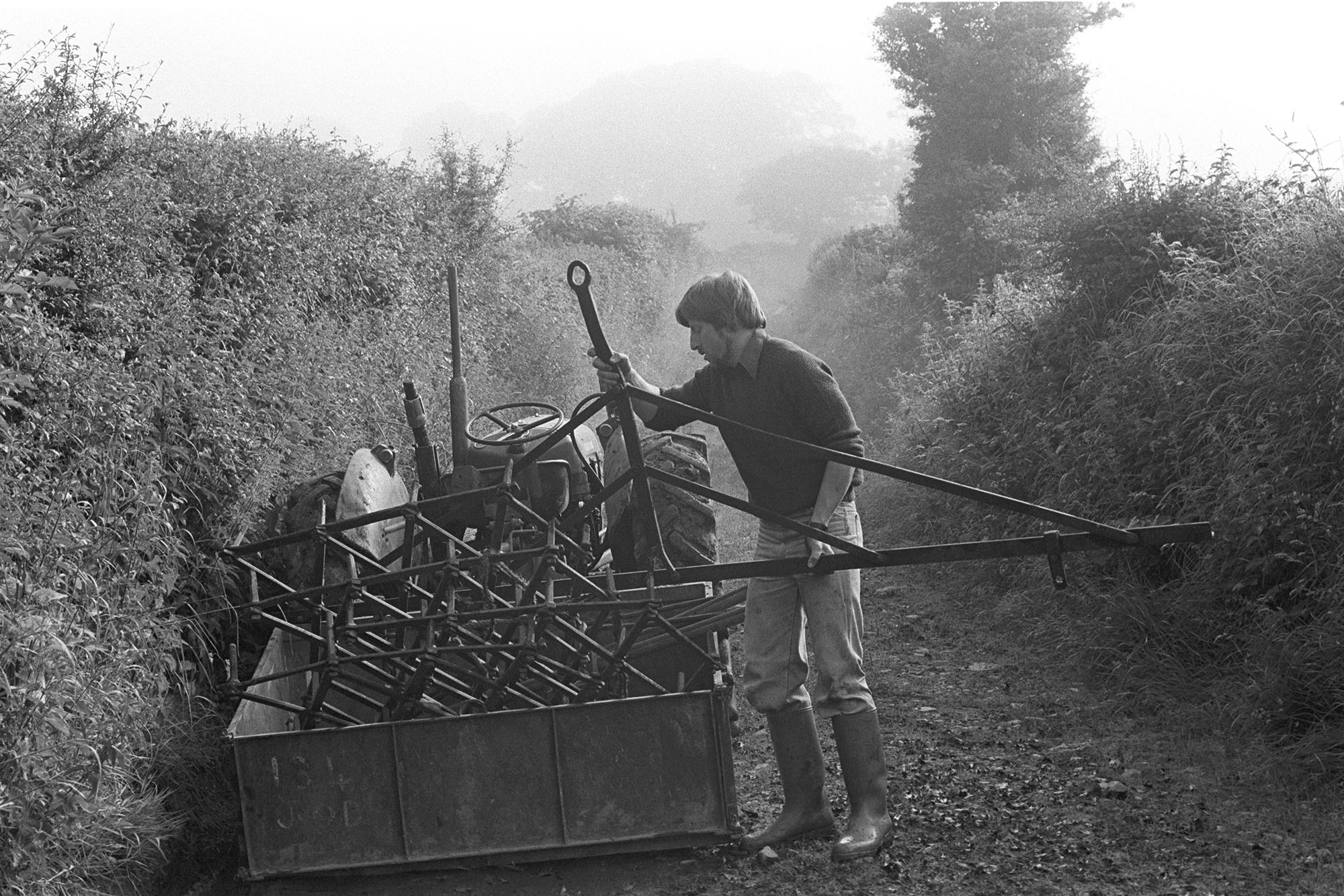 Farmer loading harrow on to tractor with link box, early morning mist. 
[Simon Berry loading a harrow into a link box attached to a tractor, in a lane at South Harepath, Beaford. The lane is covered with early morning mist.]