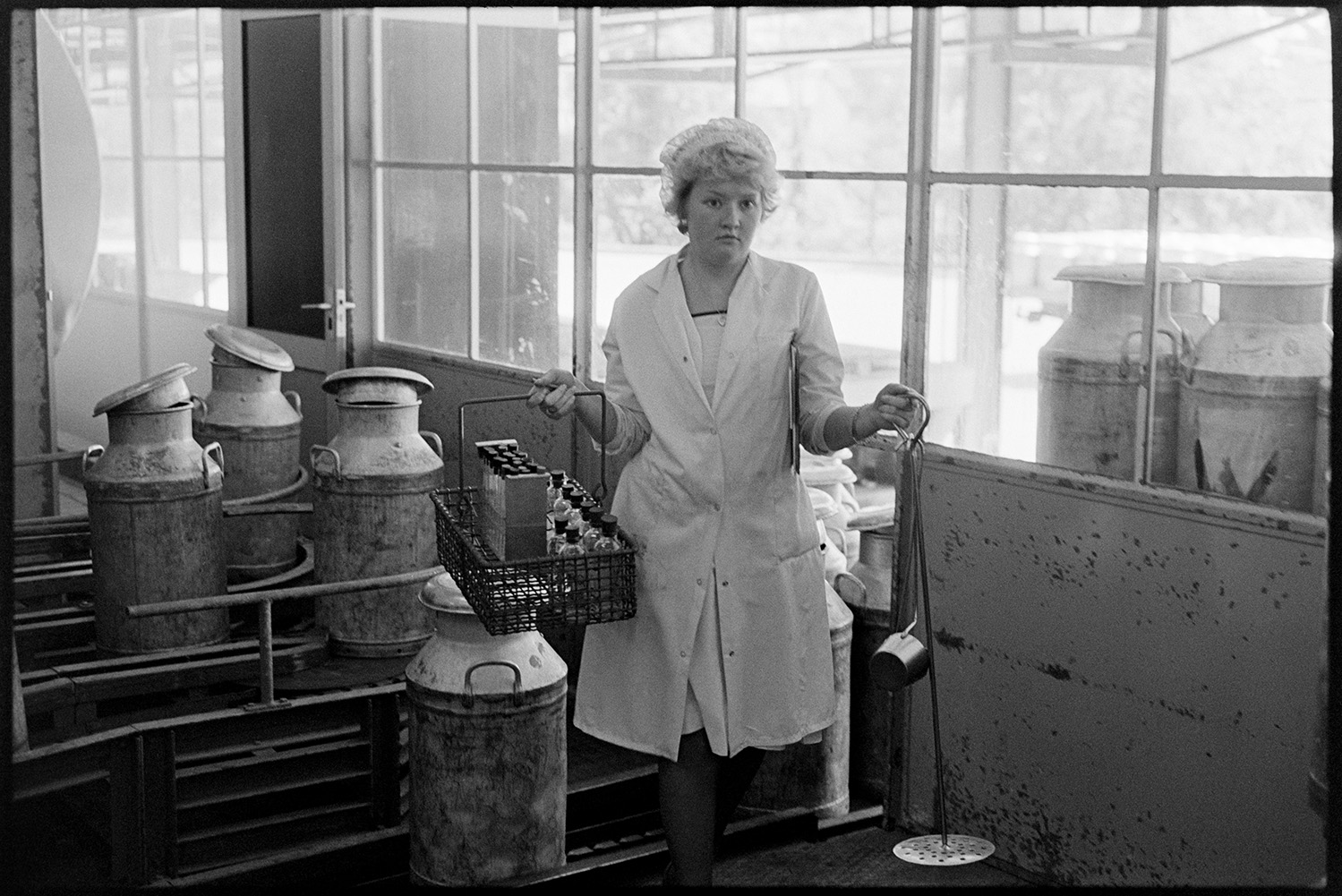 Milk arriving in churns at milk factory, sniffer checking milk, woman scientist sampling. 
[A woman carrying a crate with sample bottles and metal tools to test milk at Unigate Dairy in Torrington. Milk churns can be seen in the background of the factory.]
