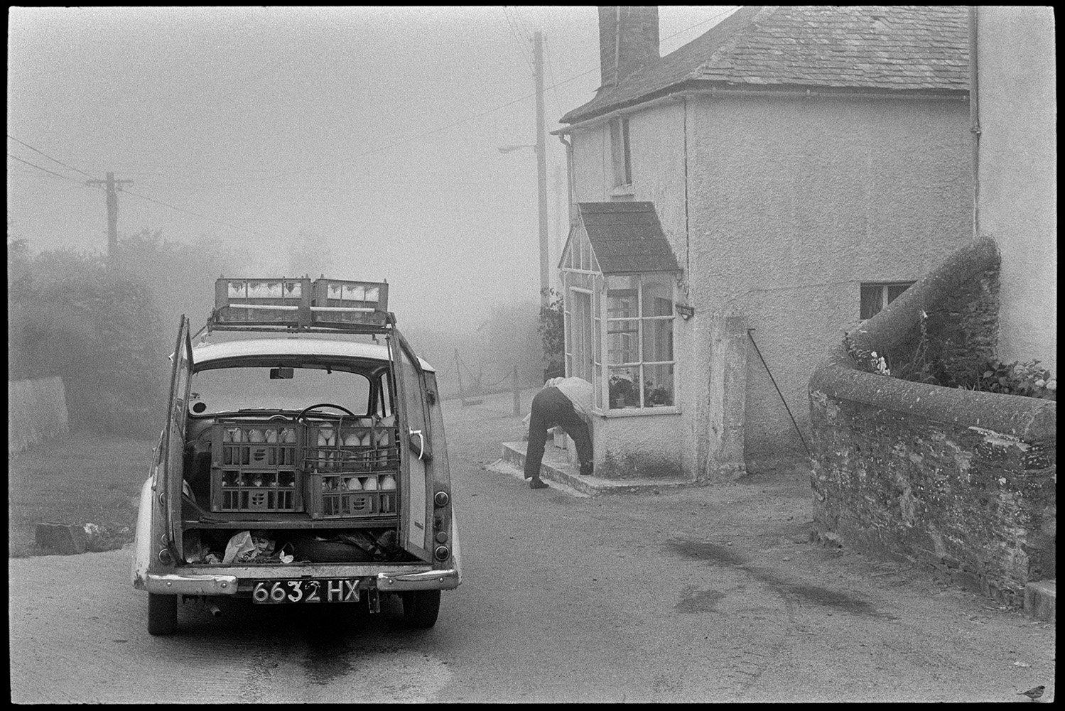Man and woman delivering milk in village from back of van, car. 
[Sid Hiscock delivering milk bottles to Rose Cottage in West Lane, Dolton, on a misty morning. His van with crates of milk bottles is parked outside the cottage.]