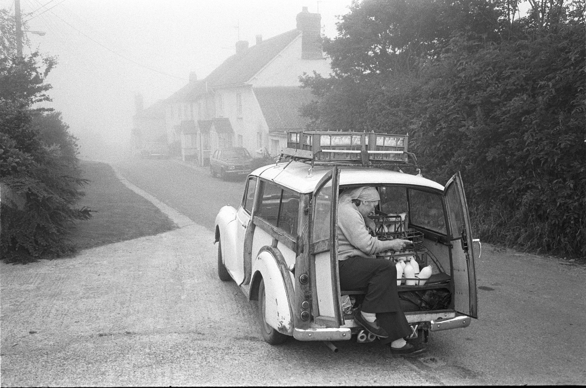 Woman checking milk deliveries in back of car in misty street. 
[Dorothy Hiscock checking milk bottles in the back of her car on her milk round in West lane, Dolton. There are more bottles secured to the roof of the car. The street is shrouded in mist.]