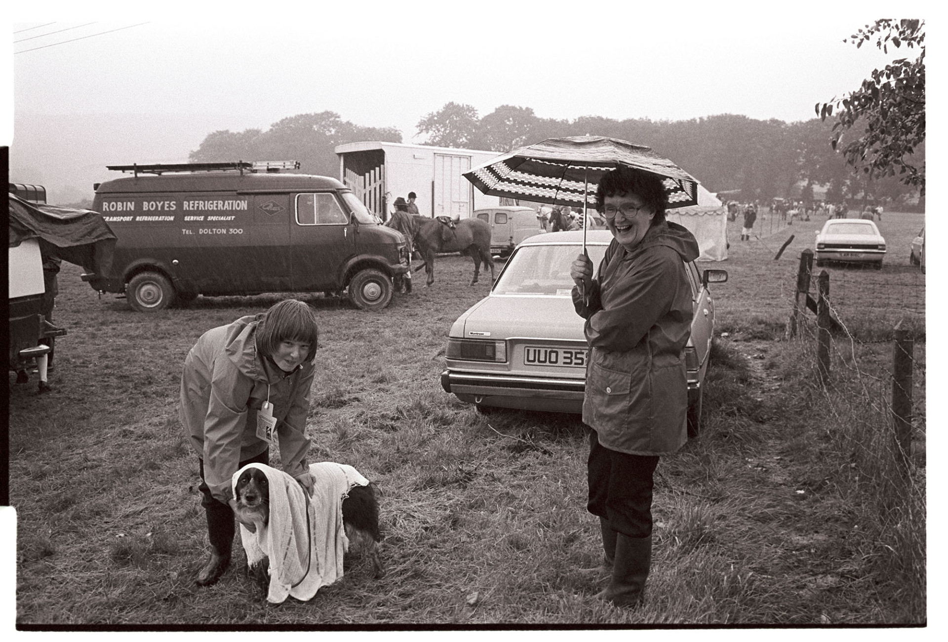 Gymkhana, dog show, trying to dry out dog in pouring rain! Humorous. 
[Two women at Beaford Gymkhana. One woman is drying off a dog with a towel and the other is standing watching under an umbrella. In the background parked cars, horse boxes, horses and riders can be seen.]
