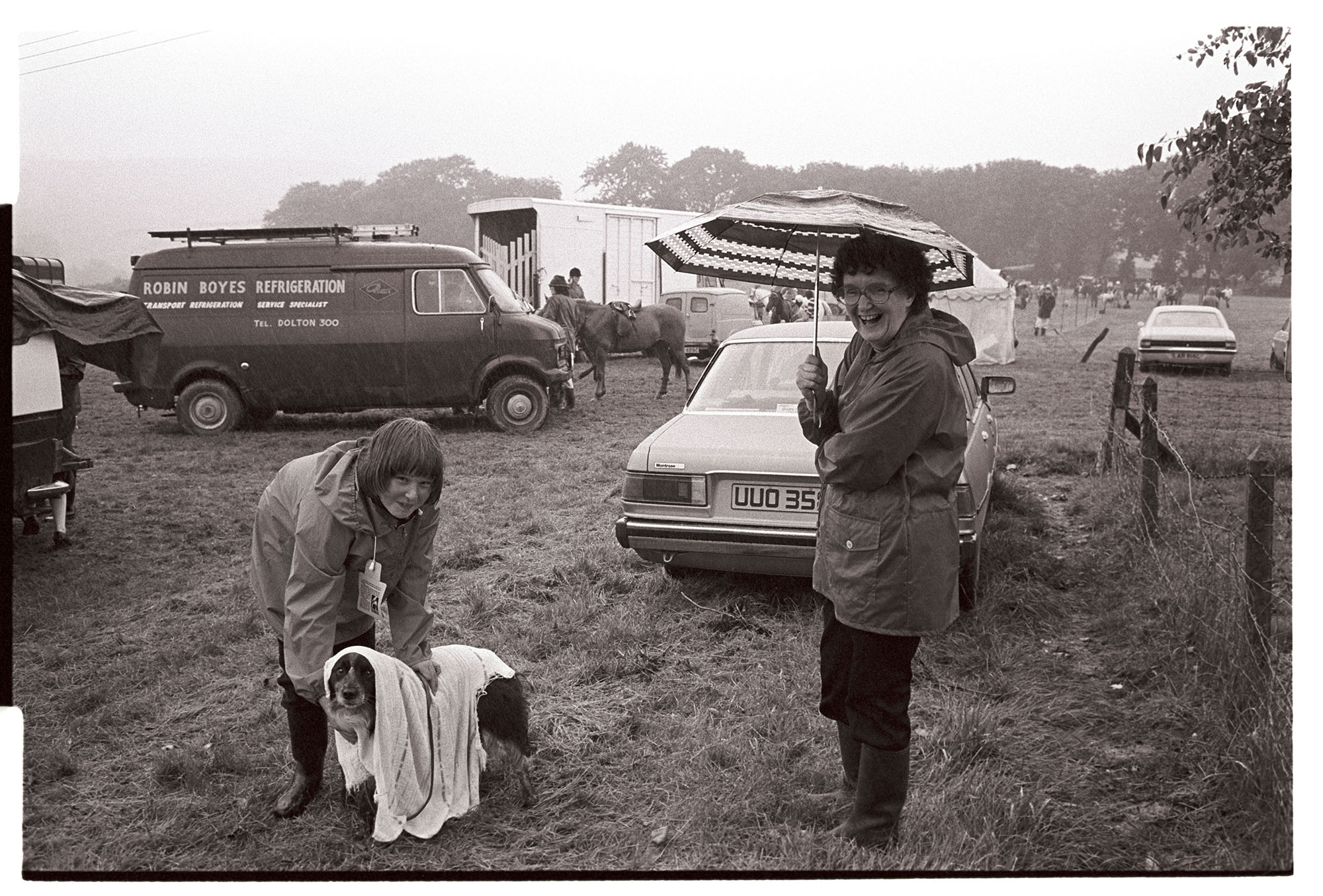 Gymkhana, dog show, trying to dry out dog in pouring rain! Humorous. <br /> [Two women at Beaford Gymkhana. One woman is drying off a dog with a towel and the other is standing watching under an umbrella. In the background parked cars, horse boxes, horses and riders can be seen.]