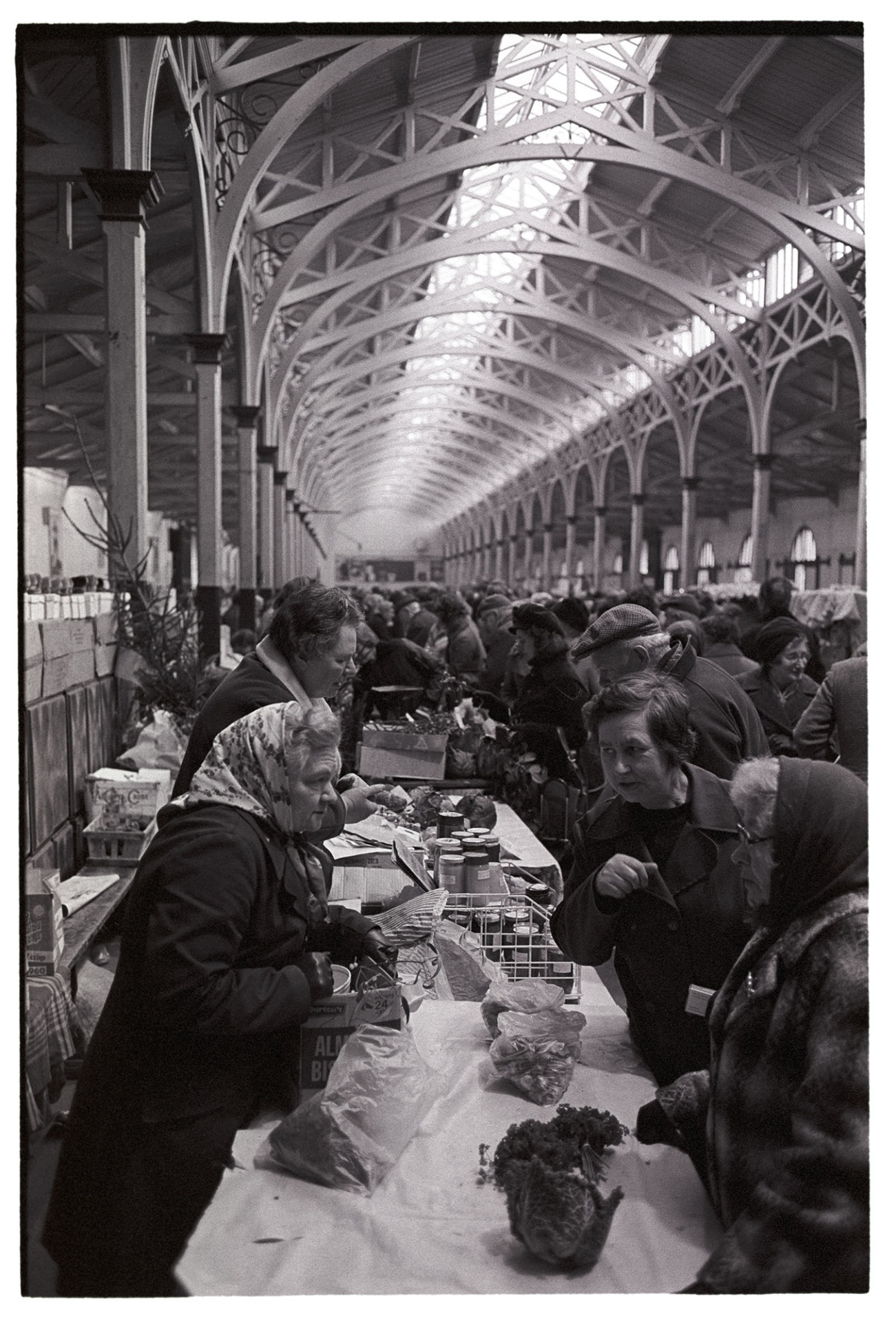 Stall at Pannier Market, women chatting. 
[Three women talking over a stall with vegetables at Barnstaple Pannier Market. Other shoppers and a stall selling jams are visible in the background.]