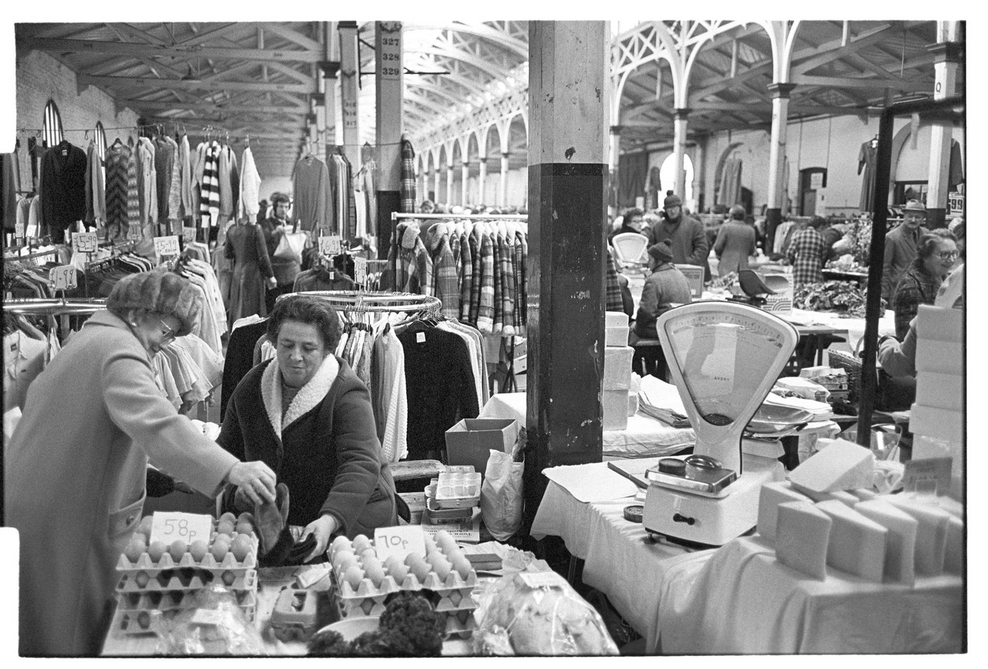 Stalls at Pannier Market. Eggs and cheese, woman serving. 
[An egg stall and cheese stall at Barnstaple Pannier Market. A woman is serving another woman on the egg stall. In the background rails of clothes and other shoppers can be seen.]