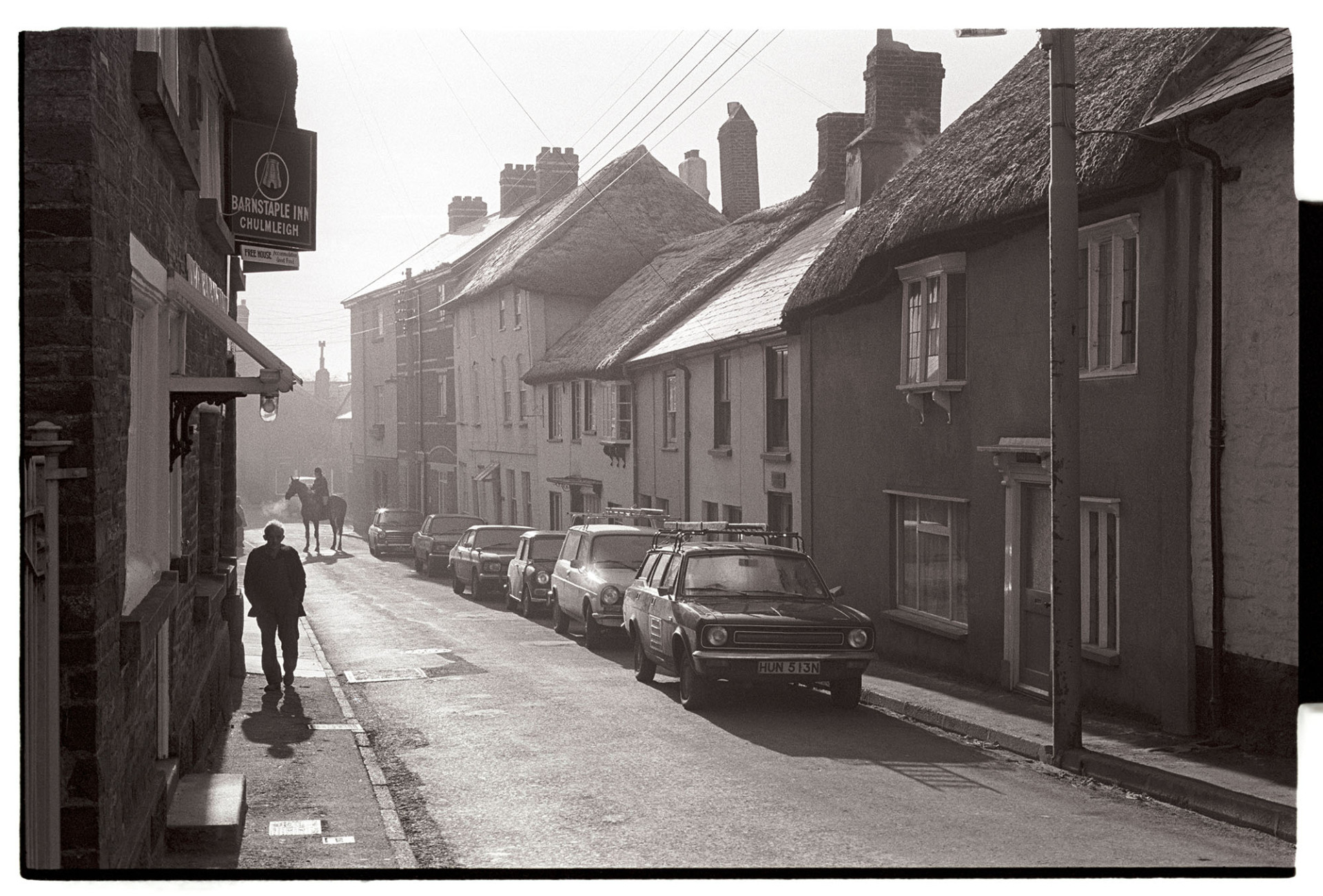 Street scene early morning with parked cars and horse rider. 
[A view of South Molton Street, Chulmleigh in the early morning. Thatched cottages, parked cars and a man walking past the Barnstaple Inn can be seen, along with a horse rider further down the street.]