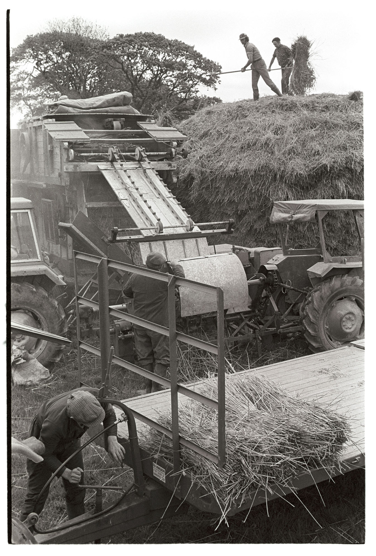 Reed comber, machine, wheat ricks, nitches loaded on trailer. 
[Two men stood on top of a wheat rick loading reed onto a reed comber attached to two tractors at Westacott Barton, Riddlecombe. Dudley Middleton is stood on the right. There is a trailer being loaded with nitches in the foreground.]