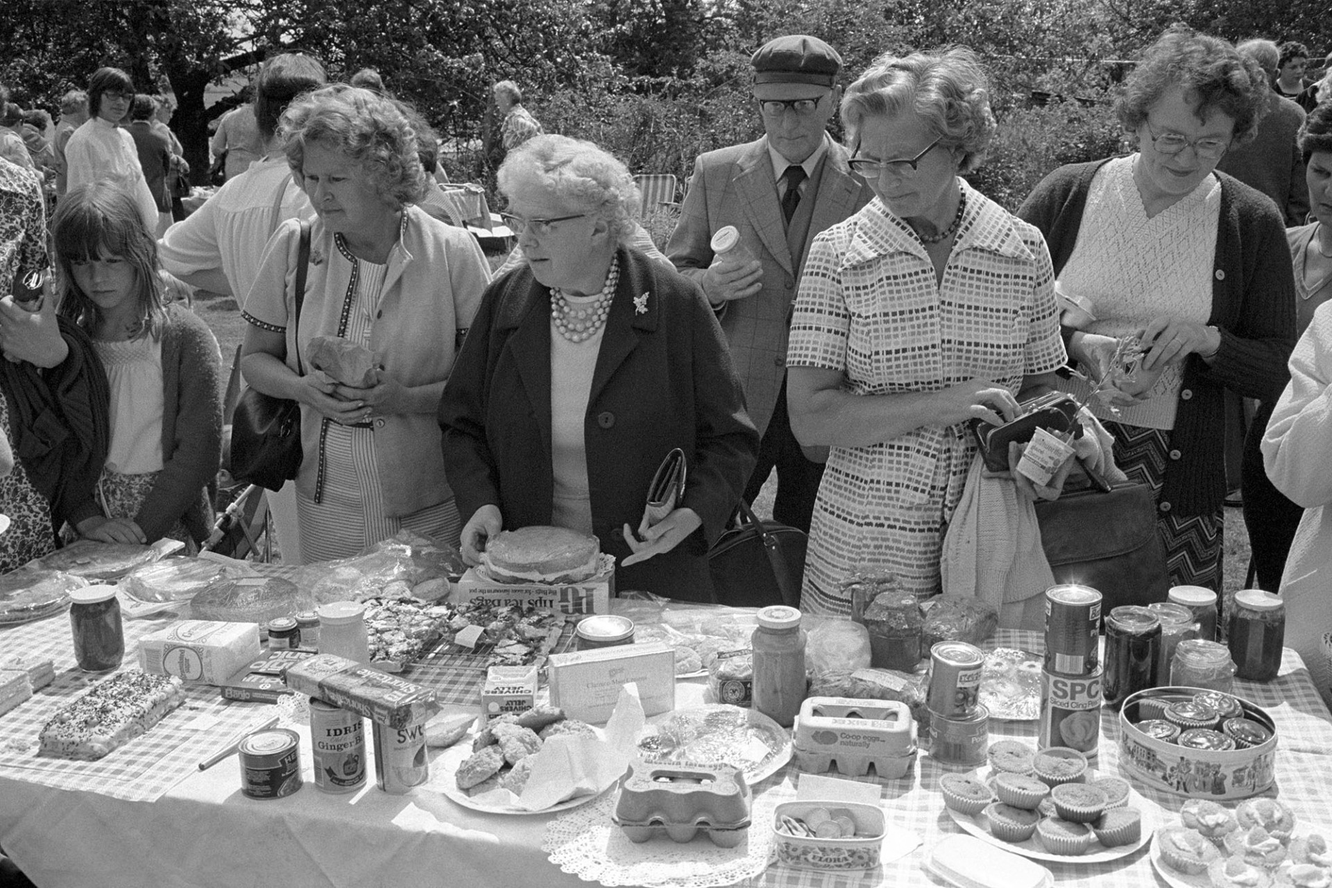 Garden party with stall and women looking at produce on table. 
[Women, a young girl and a gentleman looking at produce and groceries on a table at a garden party at Barlands, Dolton. The stall includes cakes, eggs, jars of preserves, tinned food, a box of PG tips, and biscuits.]