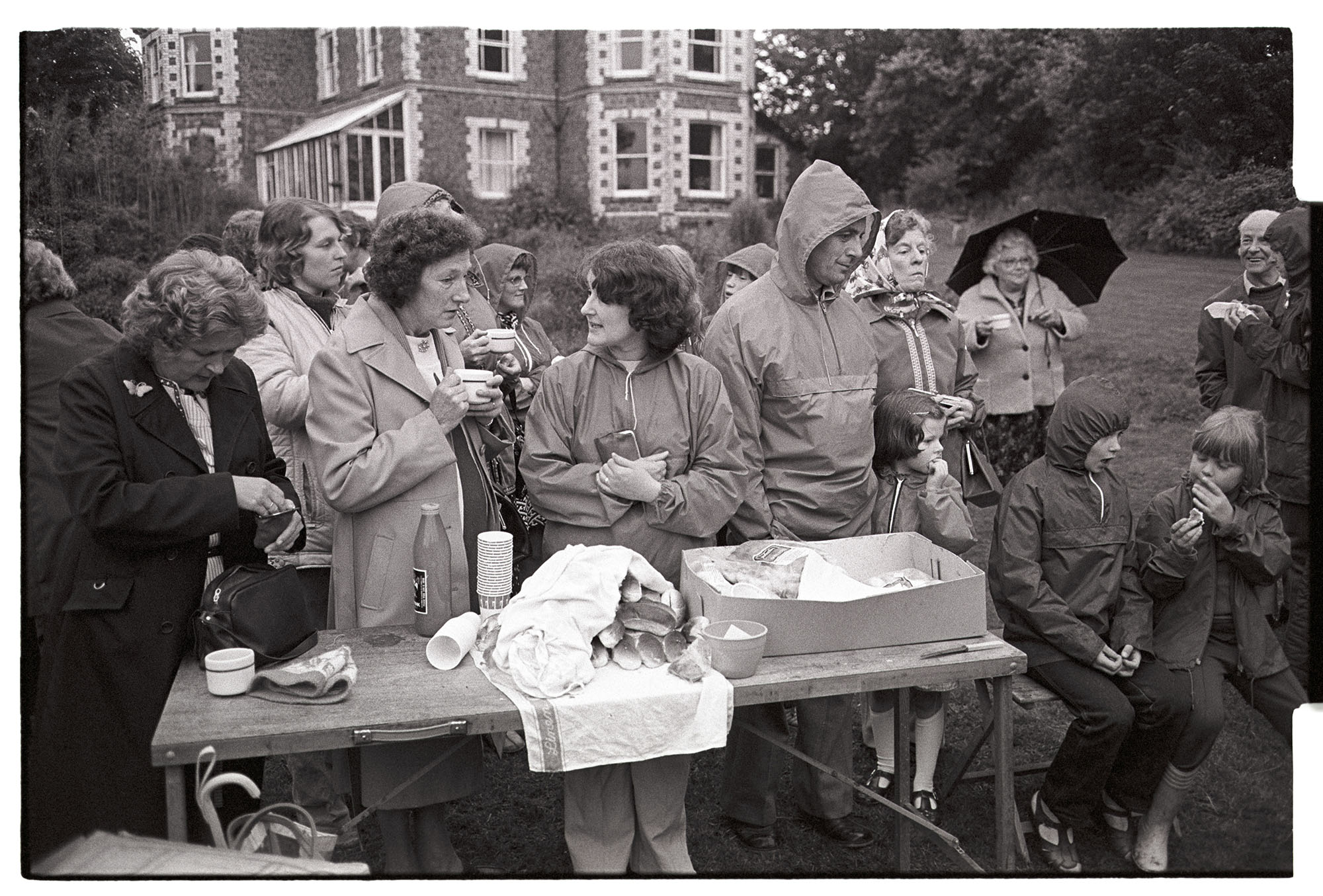 Baptist Garden Party in rain barbeque, hot dogs and tea. 
[A group of people in coats, anoraks and with umbrellas, standing in the garden by a table of hot dogs and drinking tea at the Baptist Church garden party at Barlands, Dolton. A large house is in the background.]