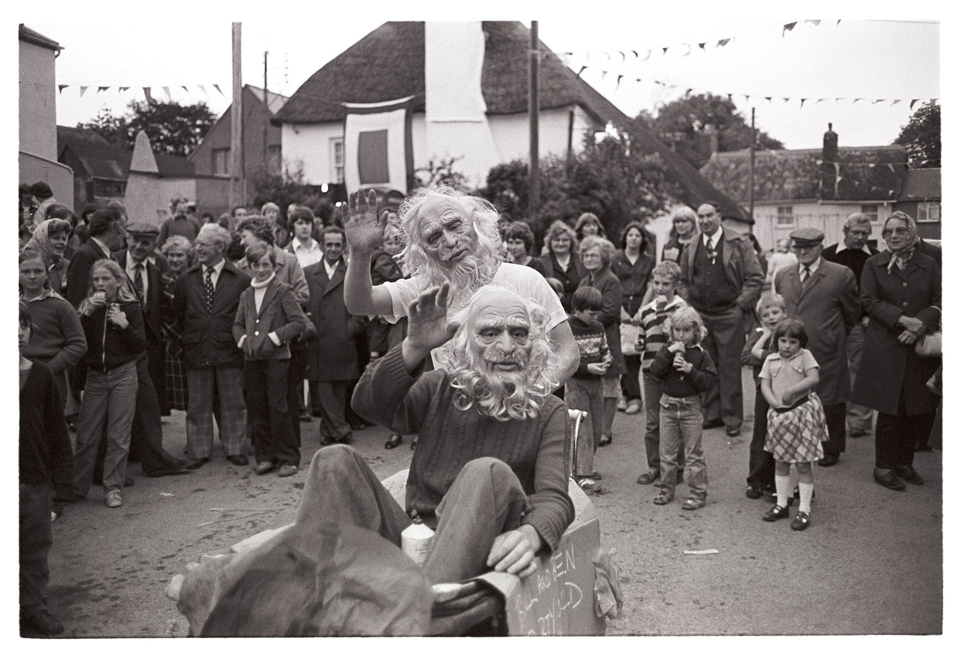 Village Fair, fancy dress, boys in masks in pram. 
[Two boys in masks, one in a pram, in a street  at Winkleigh Fair, with a crowd of people watching them, and cottages and bunting in the background.]