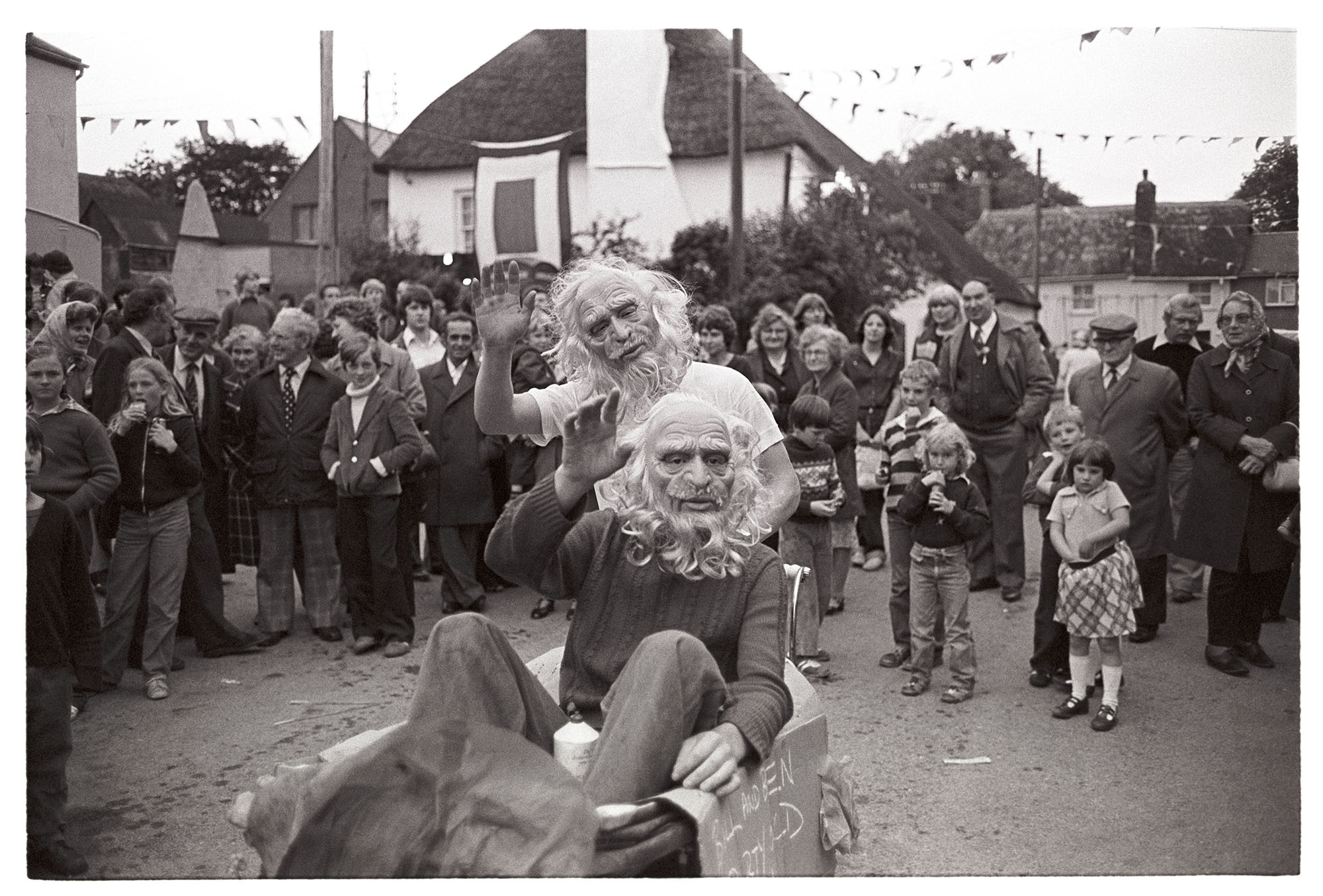 Village Fair, fancy dress, boys in masks in pram. <br /> [Two boys in masks, one in a pram, in a street at Winkleigh Fair, with a crowd of people watching them, and cottages and bunting in the background.]