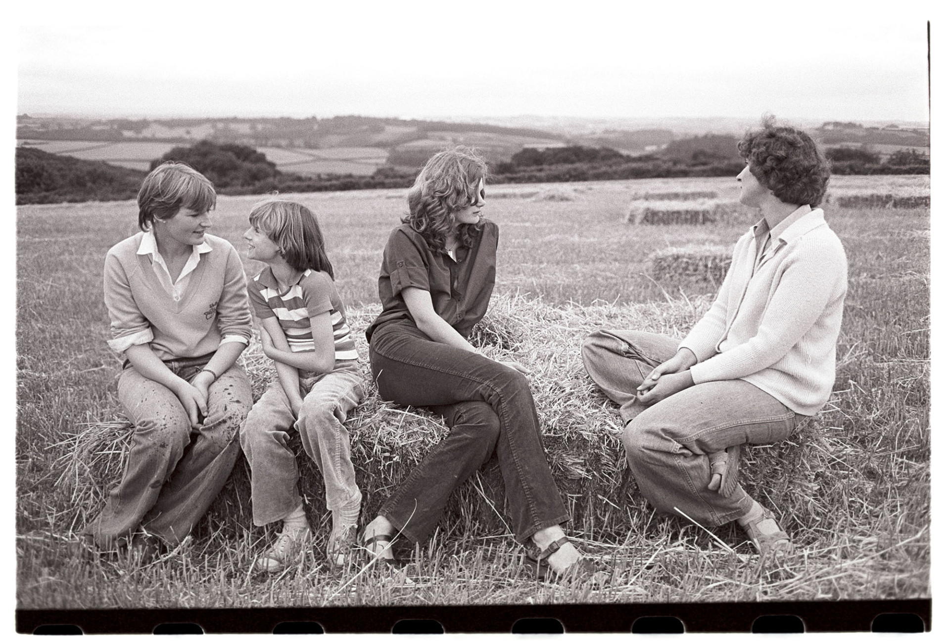 Women sitting on straw bales resting in field. 
[Kay Allin, two other women and a young girl sat on straw bales in a field at Ashwell, Dolton with a view of fields and trees in the background.]