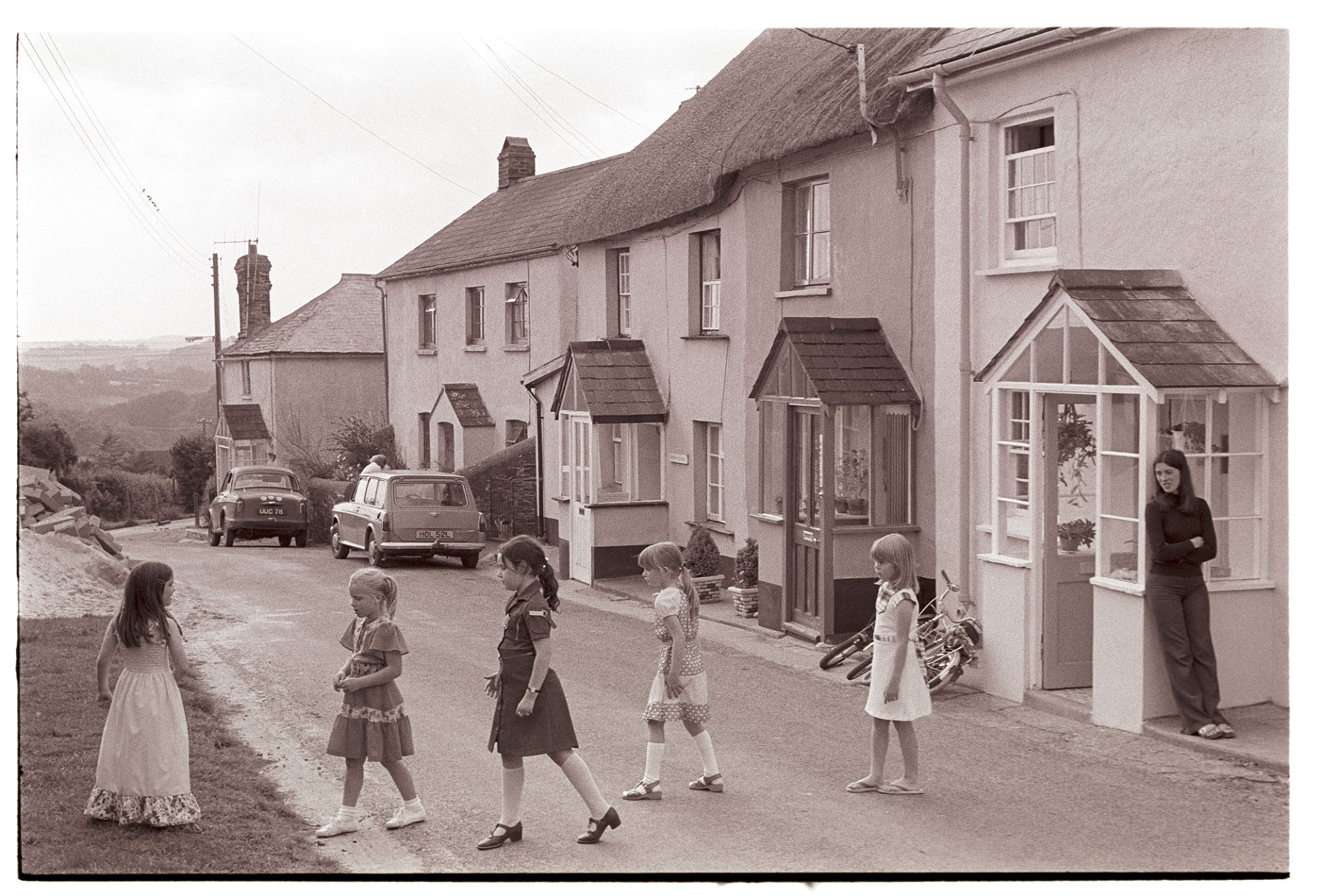 Children at birthday party playing Grandmothers Footsteps in street, party dresses. 
[A group of young girls in party dresses playing Grandmothers Footsteps in the street at West Lane, Dolton while Rosalind Squire, mother of one of the girls, looks on from the front of a house. There are more houses and parked cars down the lane in the background.]