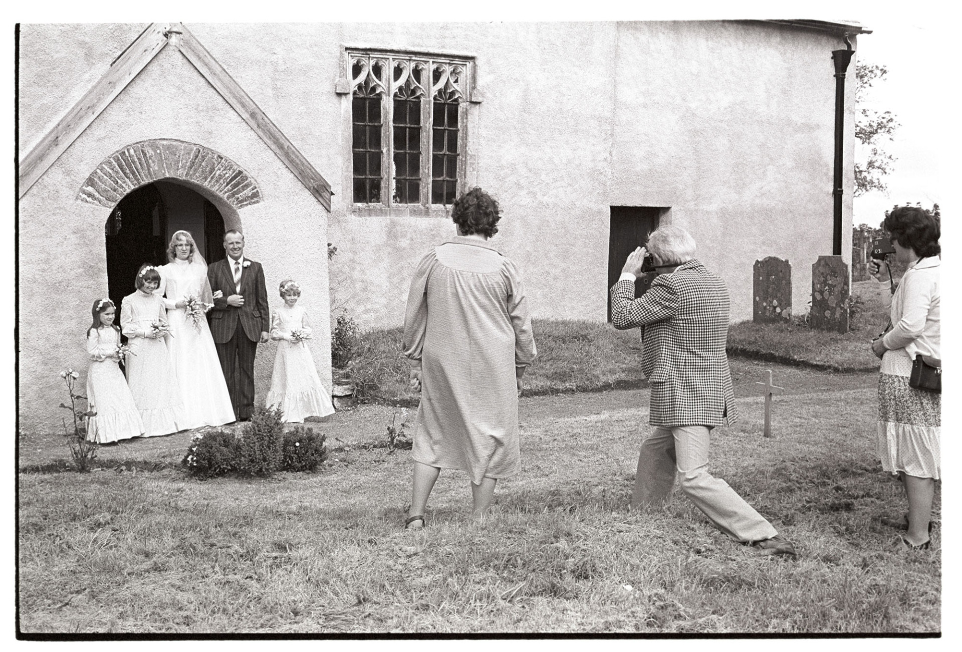 Wedding, bride and father being photographed with bridesmaids. 
[The bride, Susan Westcott, standing in the doorway of Dowland Church with her father, Stephen Squire, and three young bridesmaids. Three wedding guests are stood in the churchyard and are taking photographs.]