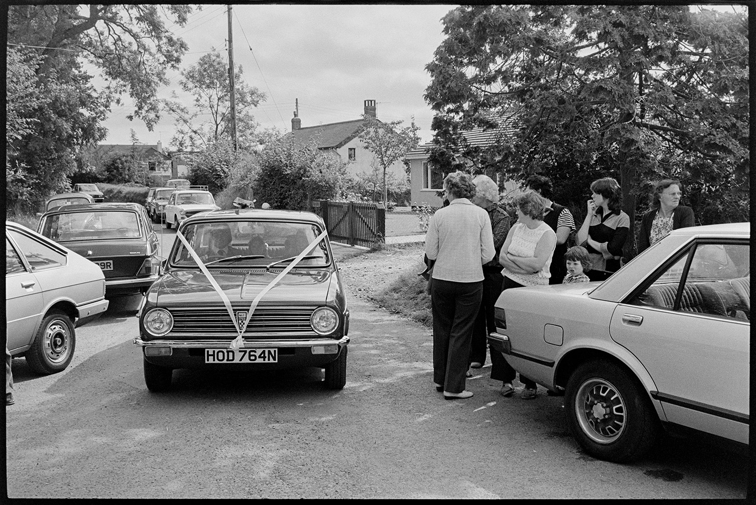 Wedding guests, bride and father arriving, photographer taking photographs, bridesmaids. 
[Onlookers watching the wedding car driving Susan Westcott and her bridesmaids along a street to Dowland Church for her wedding to Stephen Squire.]