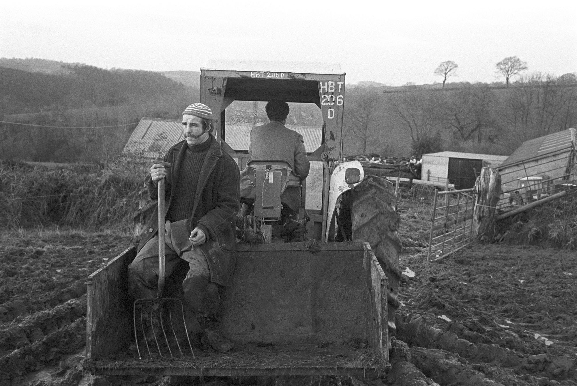 Farmer riding home sitting in link box behind tractor. 
[Roger Grigg having a lift home sitting in a link box attached to a tractor. He is holding a large fork which he has been using to spread manure in a field at Tockley, Dolton. Farm buildings are visible in the background.]