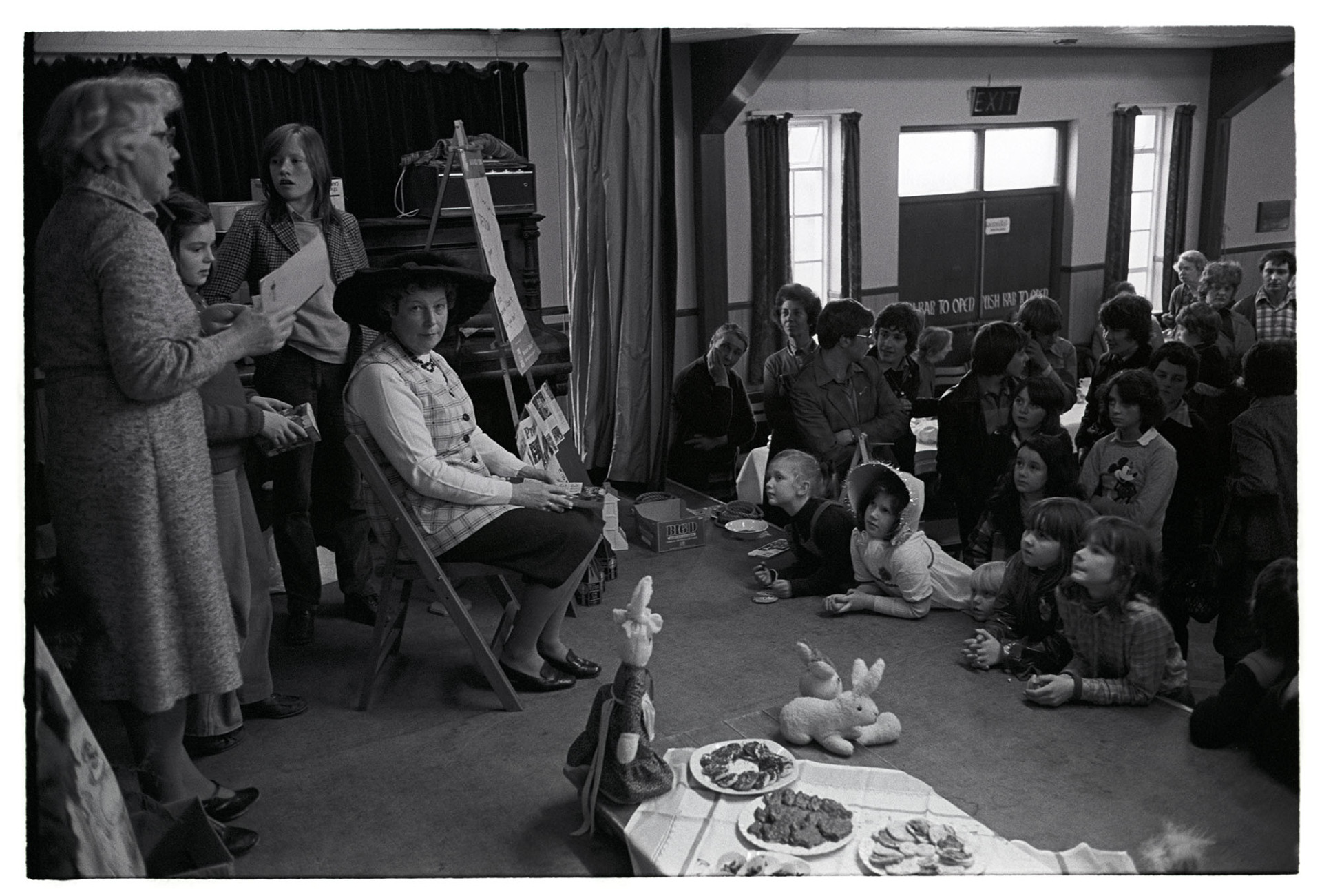 Easter party, in village hall the Easter bonnet bunnies, rabbits, hats. 
[Ethel Turner sat down in front of a group of children at an Easter party in Dolton Village Hall. Another woman is talking to the children. Toy rabbits and party food laid on a table is in front of the children.]