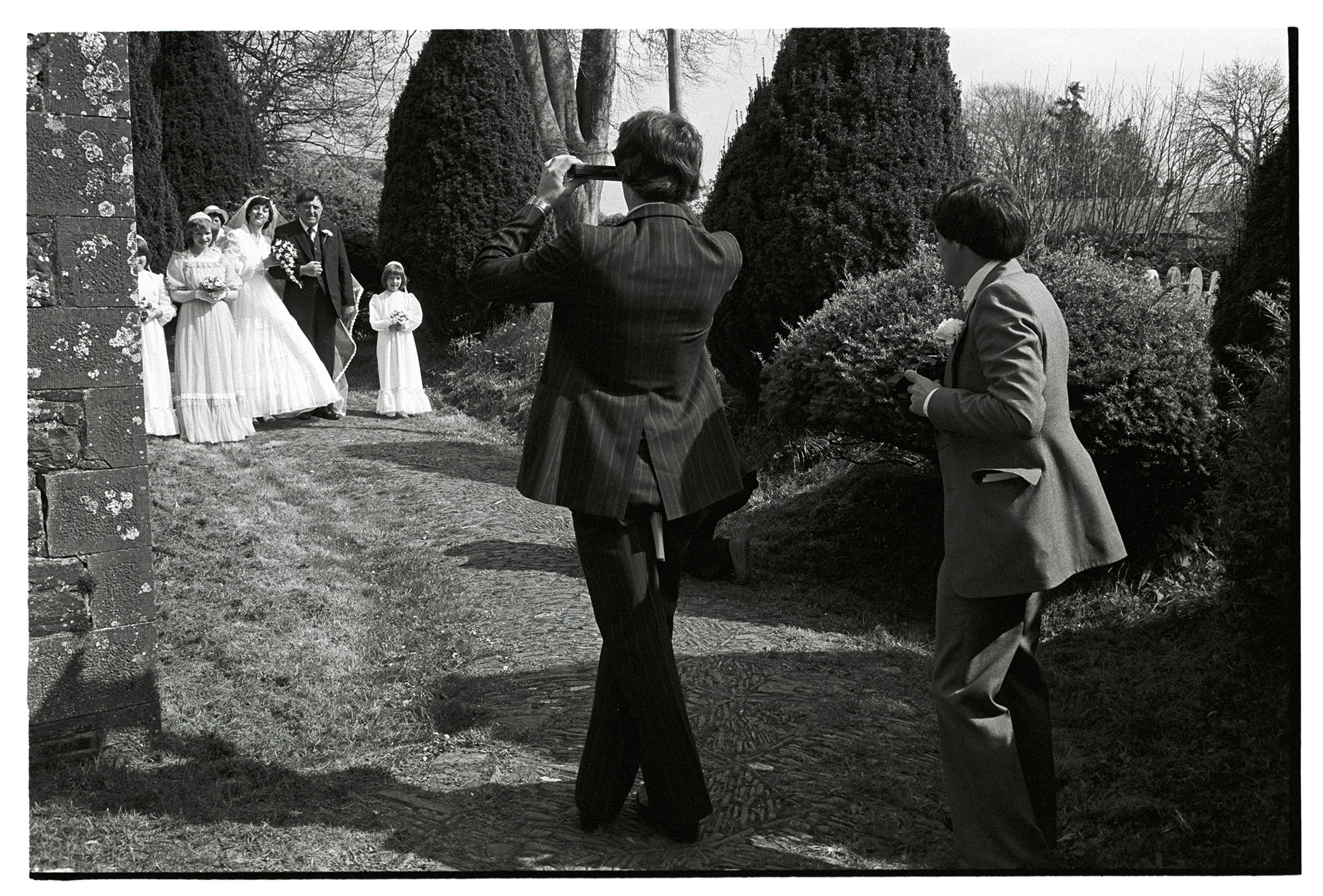 Photographing bridal group. 
[A photographer taking a picture of the bridal party at the wedding of Janet Gooch and Keith Sullivan in Merton churchyard.]
