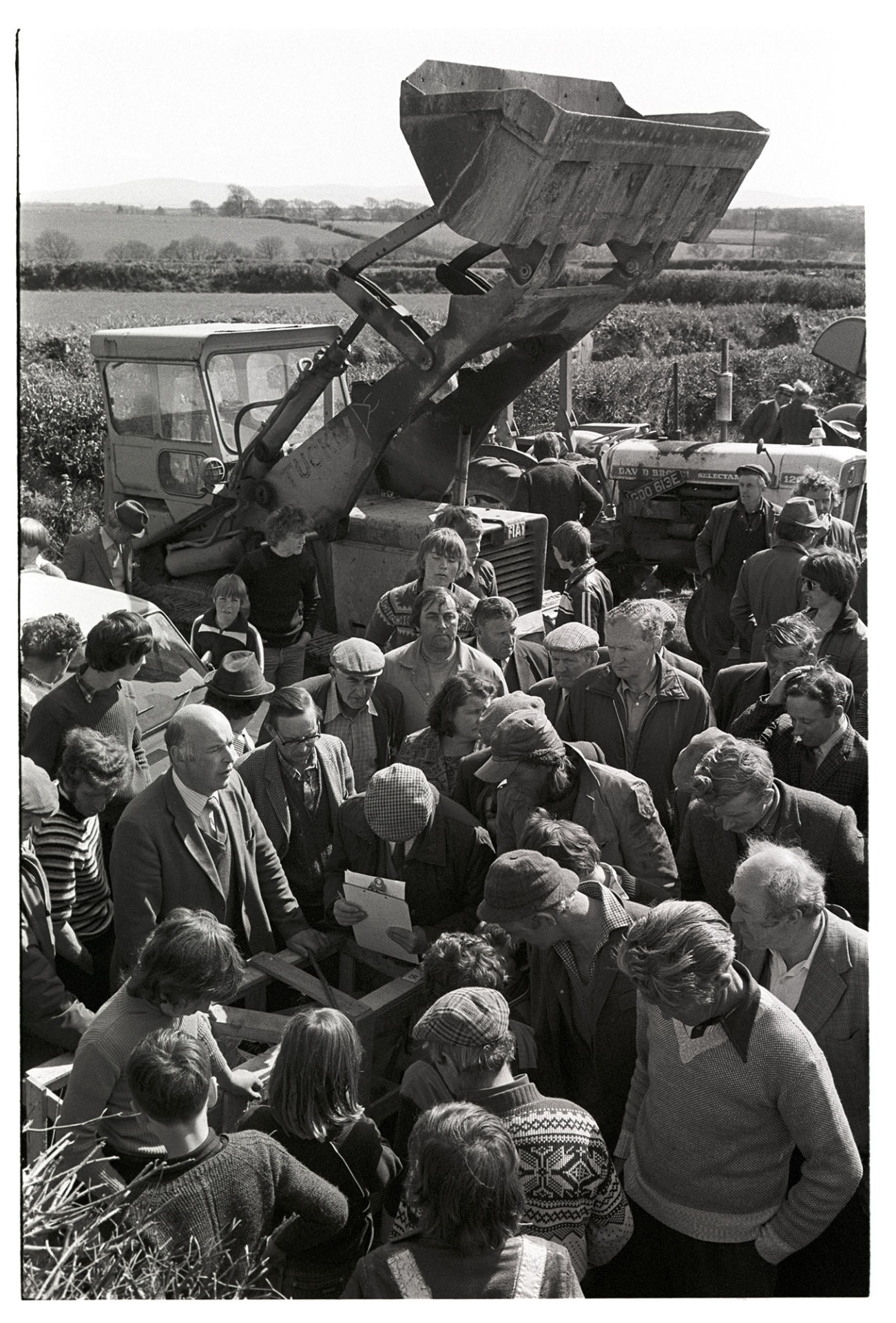Crowd at farm sale, crowds with bulldozer and auctioneer. 
[A crowd and auctioneer at a farm sale at Venton, Winkleigh. They are stood by a car, bulldozer and tractor, which are being sold.]