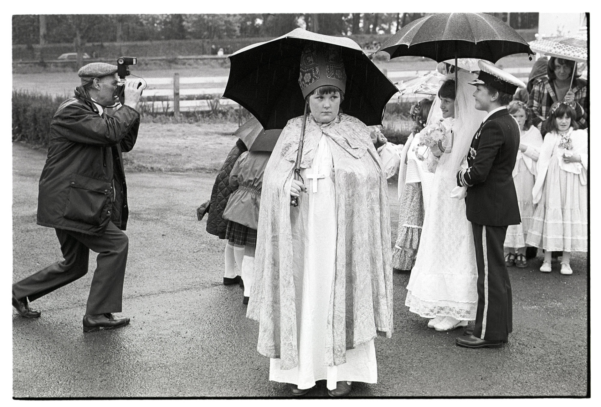 People in fancy dress being photographed, Wedding Charles and Diana, Bishop with umbrella. 
[Children in fancy dress at Torrington May Fair being photographed. Two children are dressed as Prince Charles and Lady Diana Spencer on their wedding day, and another child is dressed as a bishop. They are all holding umbrellas.]