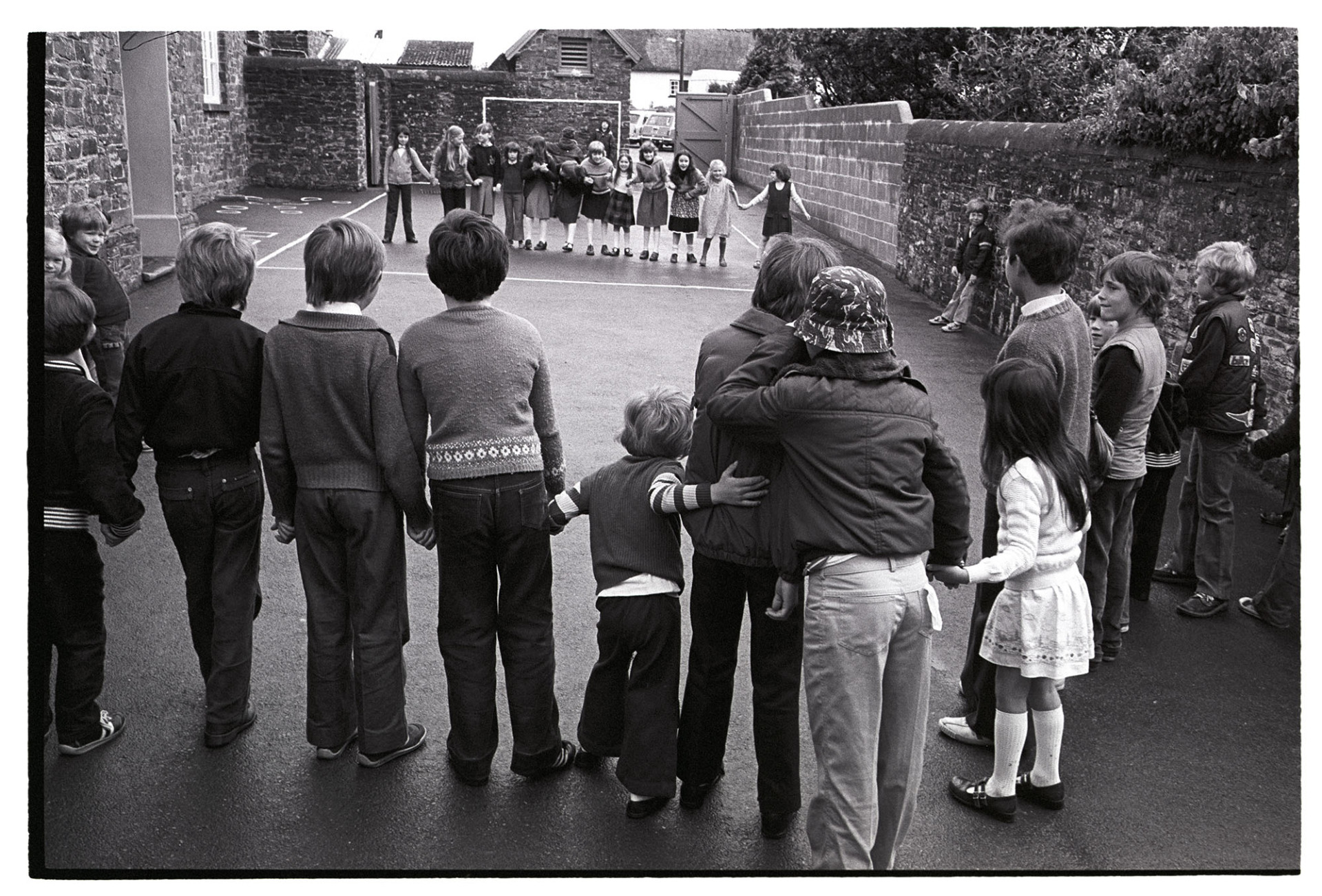 Children playing games in school playground. 
[Children playing a game in the playground of Dolton Primary School. The children are standing in two lines facing each other and holding hands.]
