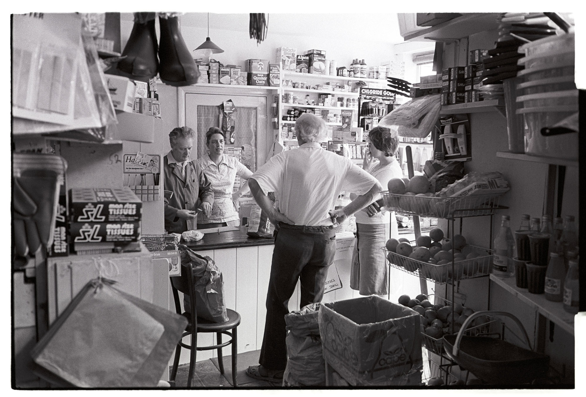 Interior of grocery stores. Customers chatting to owners. 
[Two customers talking to the owner sin Bird's Stores at Winkleigh. Various items are on display in the shop including baskets of fruit and tissues.]