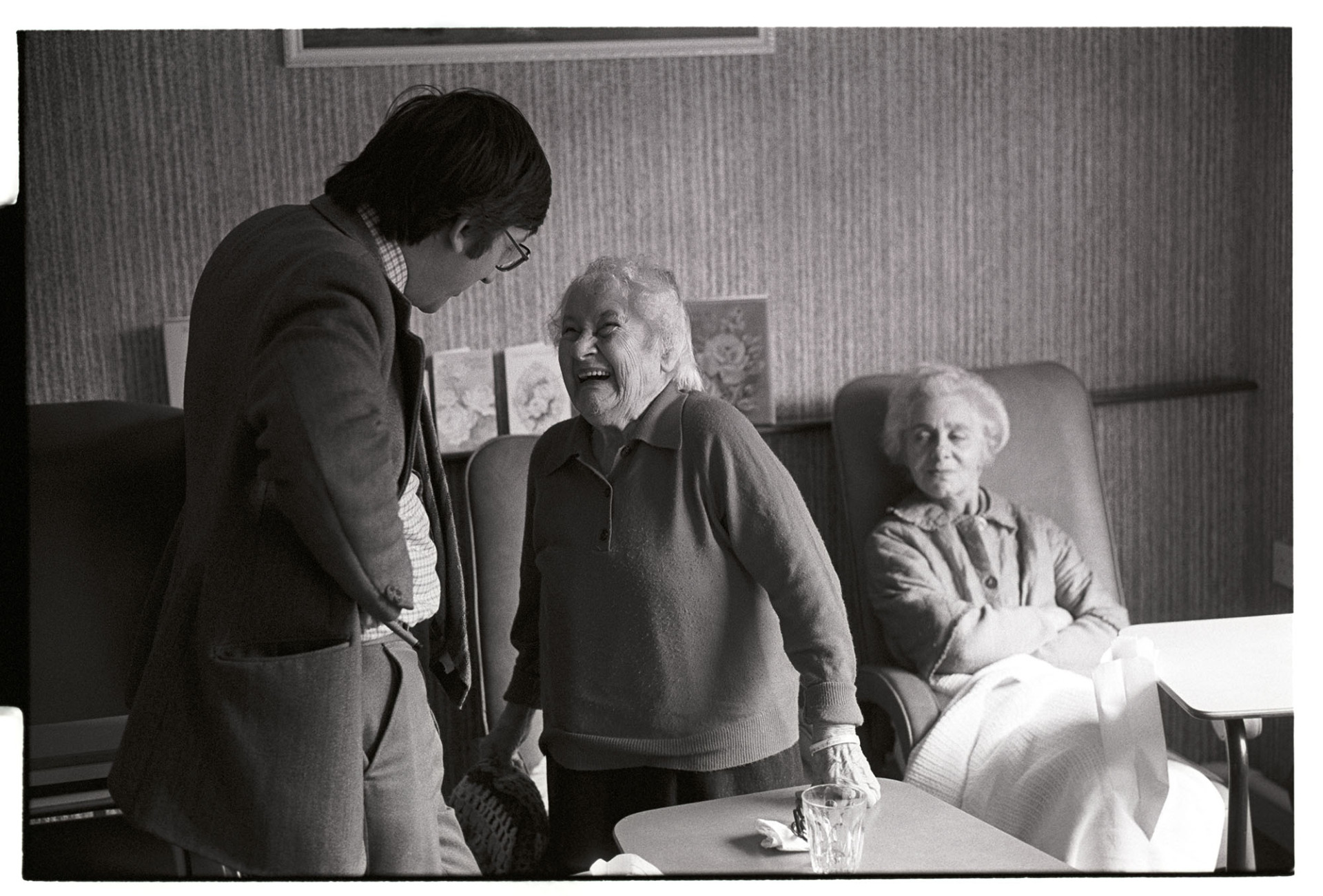 Doctor talking to elderly woman patient in day room of cottage hospital, laughing. 
[Dr Paul Bangay talking to a female patient in the day room of Torrington Cottage Hospital. The woman is laughing and another woman is sat in a chair in the background.]