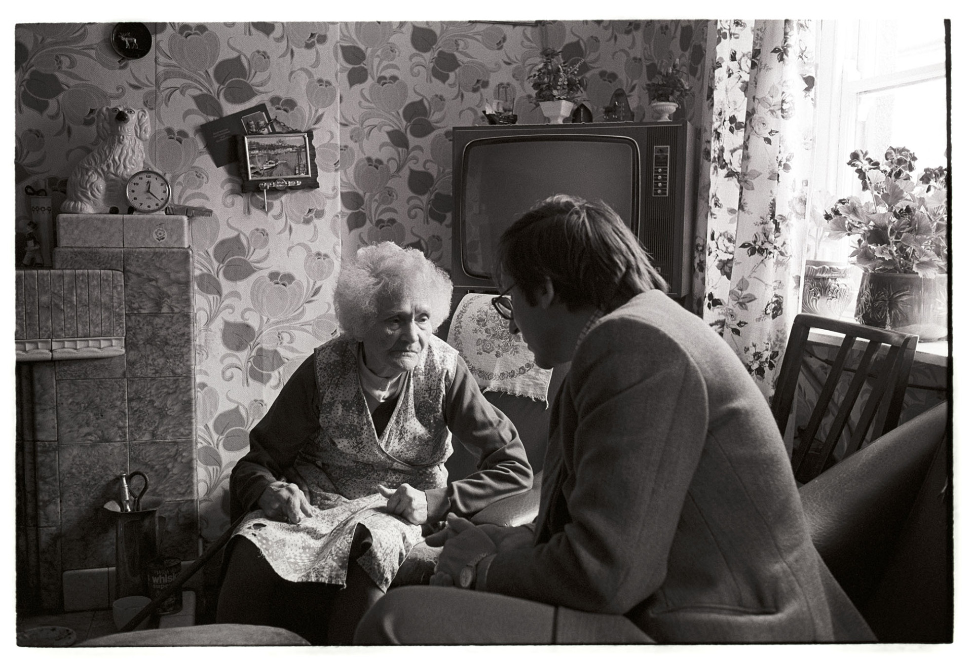 Doctor talking to elderly woman at home. 
[Dr Paul Bangay talking to a woman in her living room at her home in Torrington. A television, fireplace, ornaments on the mantelpiece and patterned wallpaper can be seen in the background.]