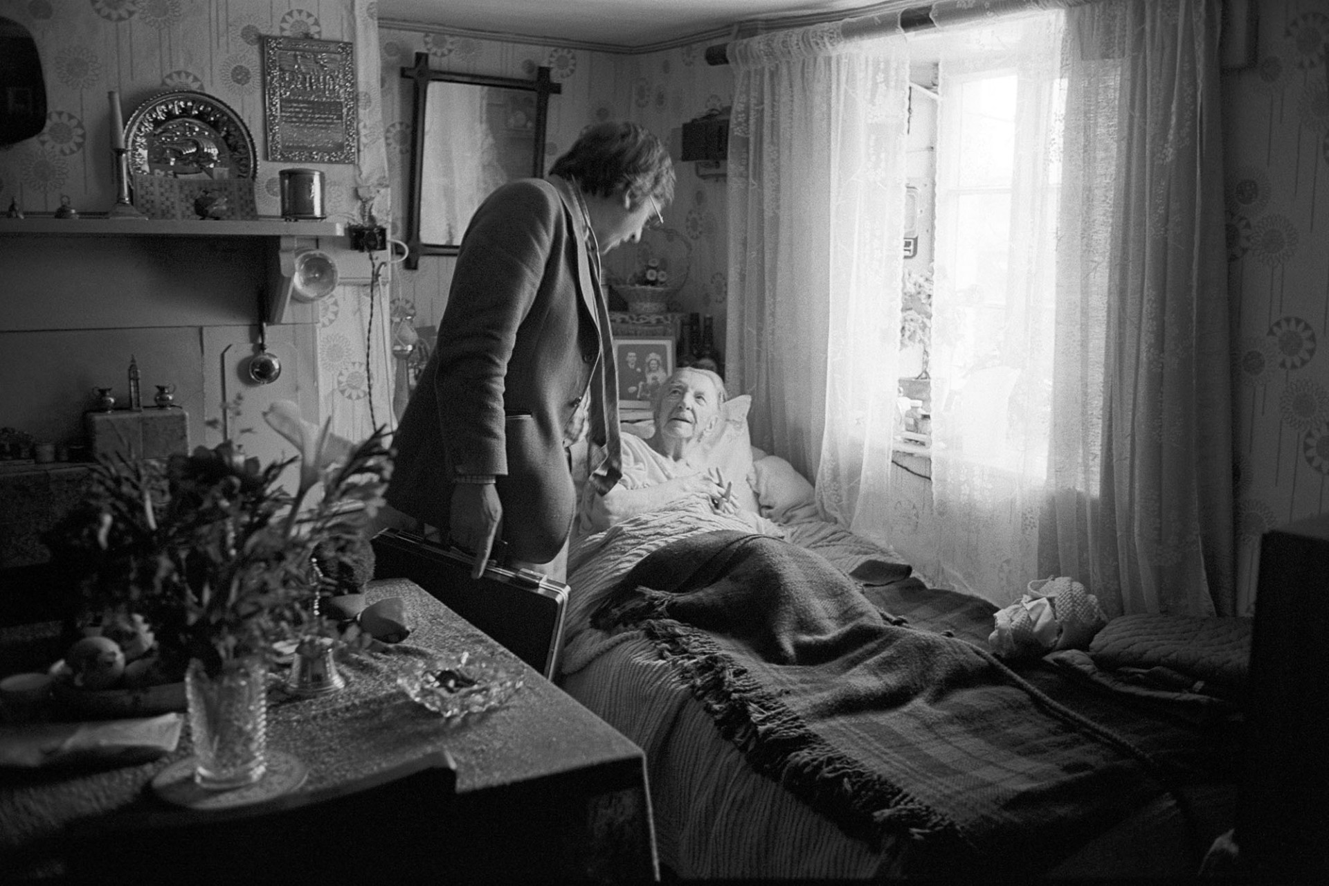 Doctor visiting elderly woman in bed, chatting. 
[Dr Paul Bangay talking to a woman in bed at her home in Langtree. Pictures and photographs are visible, as well as a vase of flowers on a table by her bed and ornaments on her mantelpiece.]
