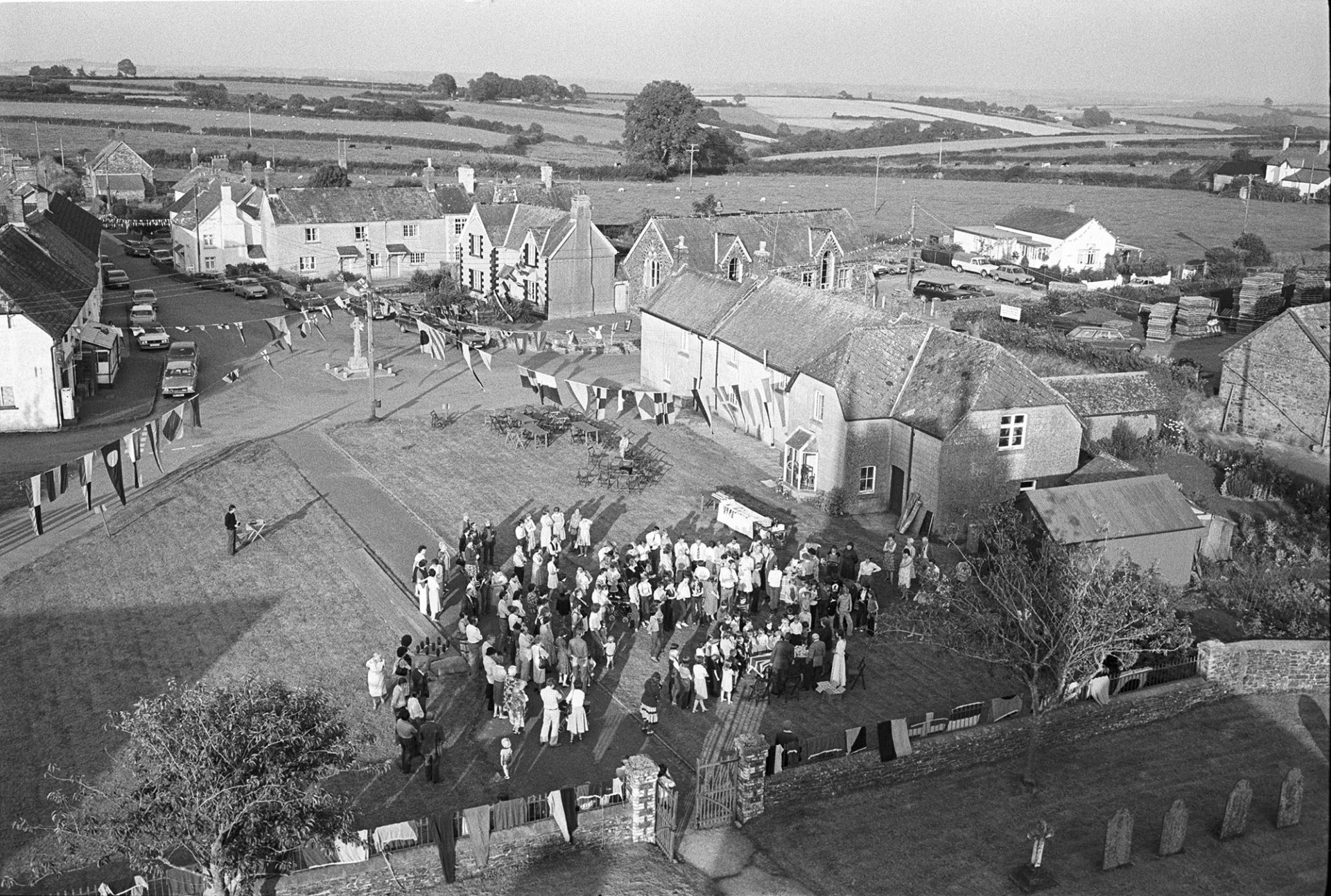 Royal Wedding. Assembling for presentation of commemorative mugs. View from church tower.
[A view taken from Ashreigney Church tower of people gathered on the green for the presentation of mugs for the Royal Wedding of Prince Charles and Lady Diana Spencer. A view of the village decorated with flags and bunting is visible behind the crowd.]