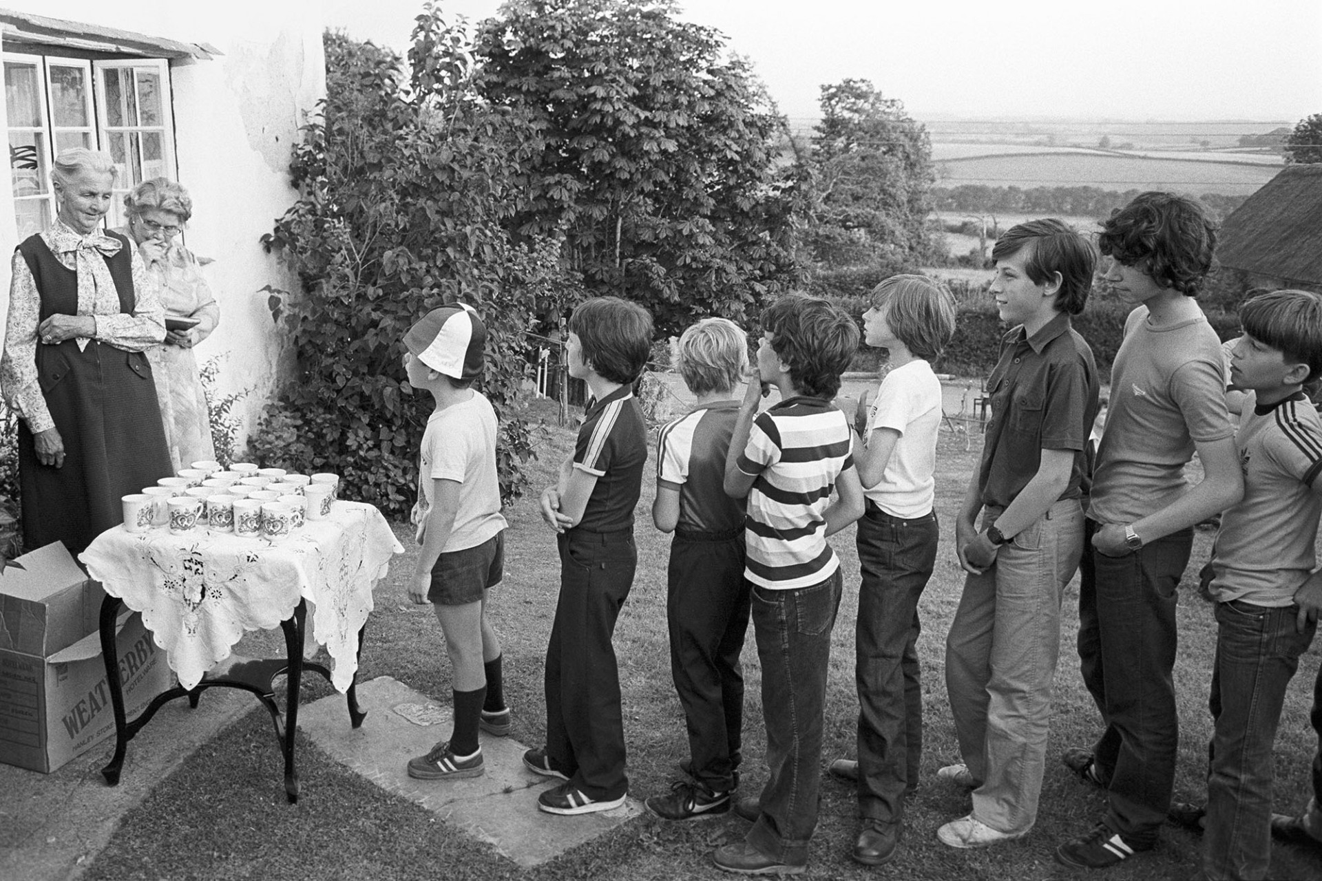 Royal Wedding. People queuing for royal mugs in front of cottage.
[Children queueing up in a garden at Iddesleigh for the presentation of mugs to commemorate the Royal Wedding of Prince Charles and Lady Diana Spencer. Two ladies are waiting to present the mugs.]