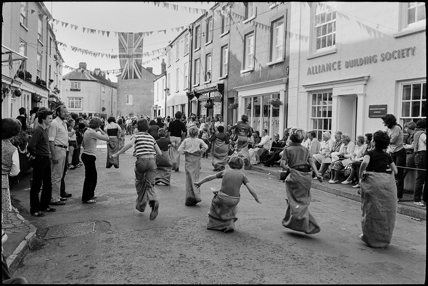 Sack race, eating buns on string in street. 
[Boys taking part in a sack race in a street at Chulmleigh Fair. They are racing past the Alliance Building Society building. People are lining the street to watch the race. Bunting and a Union Jack flag are hung across the street.]