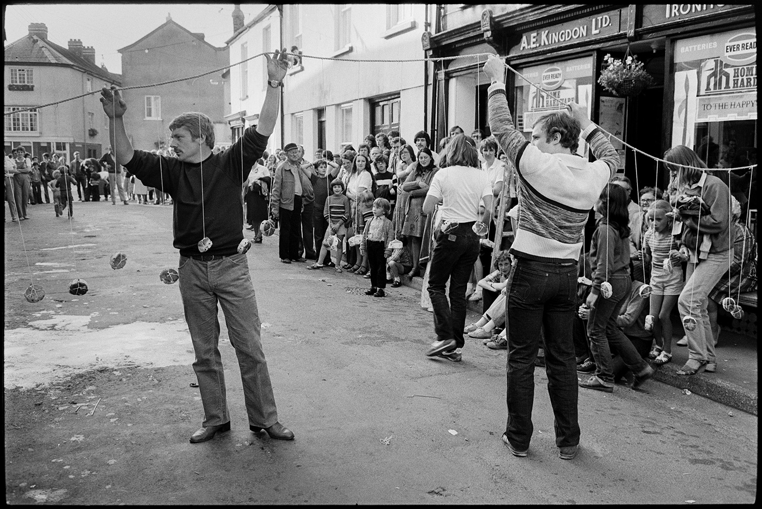 Sack race, eating buns on string in street. 
[Two men hoisting a string of buns across a street outside A E Kingdon Ironmongers shop in Chulmleigh, for children to eat in a competition at Chulmleigh Fair. People are gathered around to watch.]