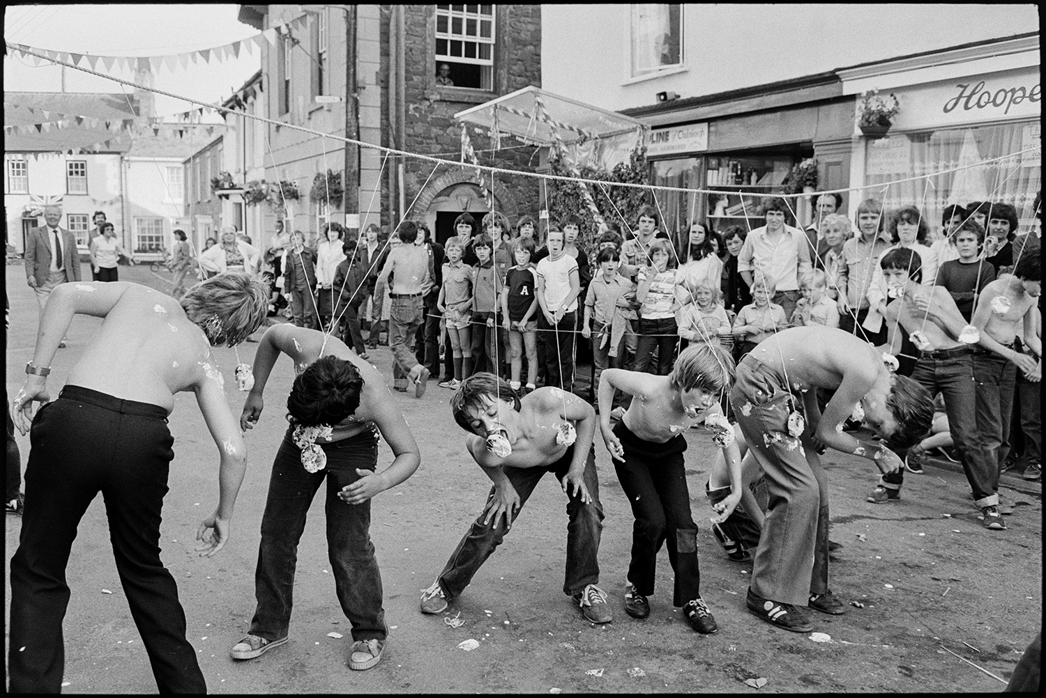 Sack race, eating buns on string in street. 
[Boys eating buns suspended on strings across a street, in a competition at Chulmleigh fair. People are gathered around watching.]