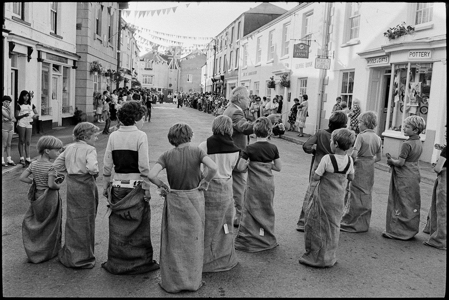 Sack race, eating buns on string in street. 
[Boys lined up ready for a sack race in a street in Chulmleigh at Chulmleigh Fair. The street is decorated with bunting and spectators are lining the pavements ready to watch the race. A sing for Lloyds bank can be seen on one of the buildings.]