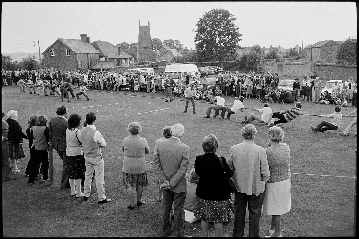 Tug o war in park, spectators. 
[People watching a tug of war match in a park at Chulmleigh Fair. Chulmleigh church tower can be seen in the background.]