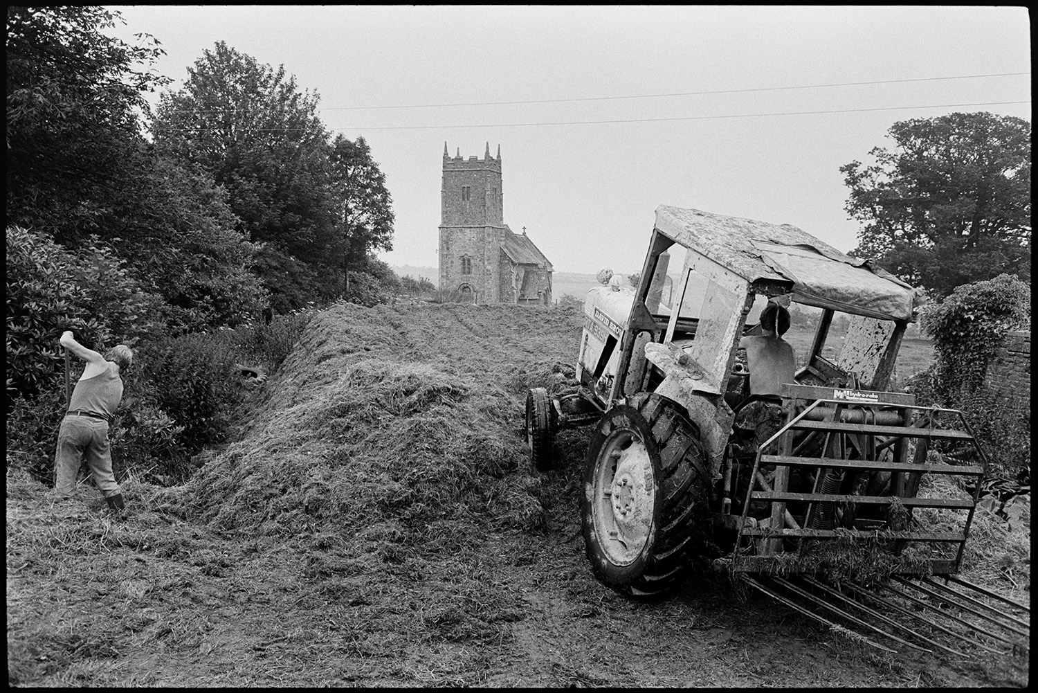 Farmers building silage clamp in front of redundant church.
[Two men with a tractor preparing a silage clamp near the redundant church at Ashbury, Northlew. One man is using a fork to dig a base for the clamp.]