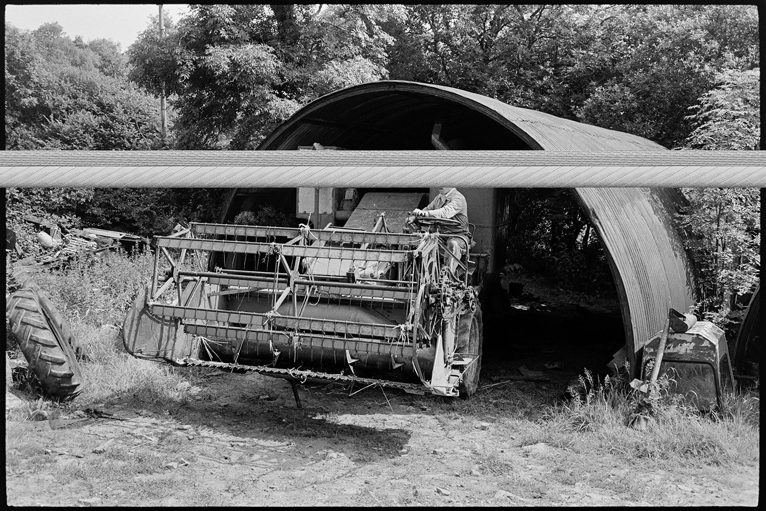 Agricultural engineers workshop, mending puncture, tractors, combine harvester. 
[A man driving a combine harvester out from under a corrugated iron arched shed, at an agricultural engineer's premises, at Hollocombe.]