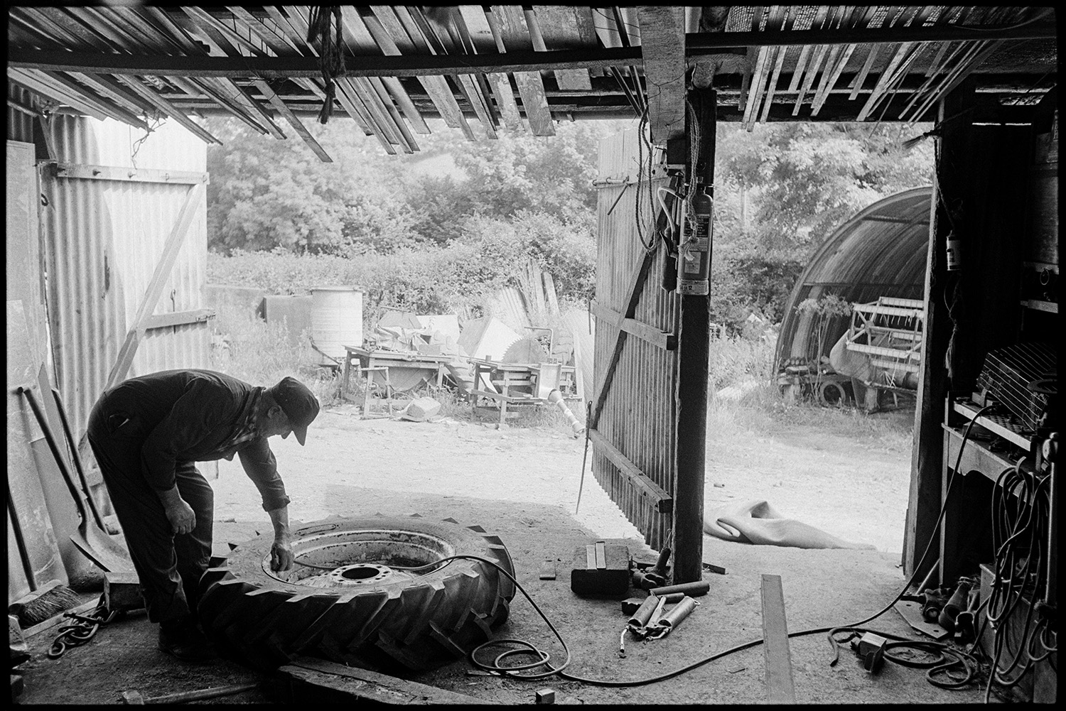 Agricultural engineers workshop, mending puncture, tractors, combine harvester. 
[A man mending a puncture in a tractor tyre in a corrugated iron shed at an agricultural engineer's workshop at Hollocombe.]