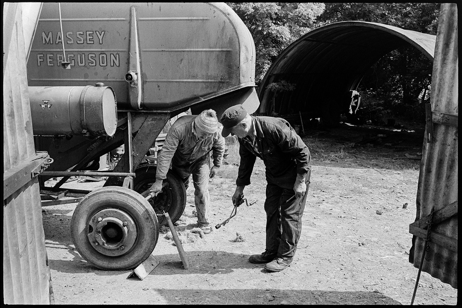 Agricultural engineers workshop, mending puncture, tractors, combine harvester. 
[Two men checking a combine harvester at an agricultural engineer's premises at Hollocombe. They are looking at the tyres.]
