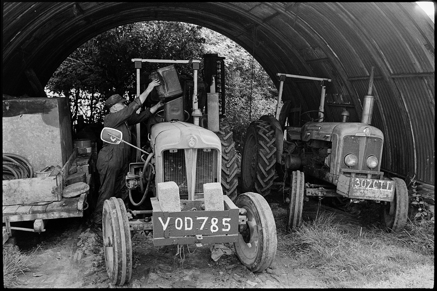 Agricultural engineers workshop, mending puncture, tractors, combine harvester. 
[A man putting fuel into a tractor under a corrugated iron arched shed at an agricultural engineer's premises in Hollocombe. Another tractor and other machinery is in the shed.]