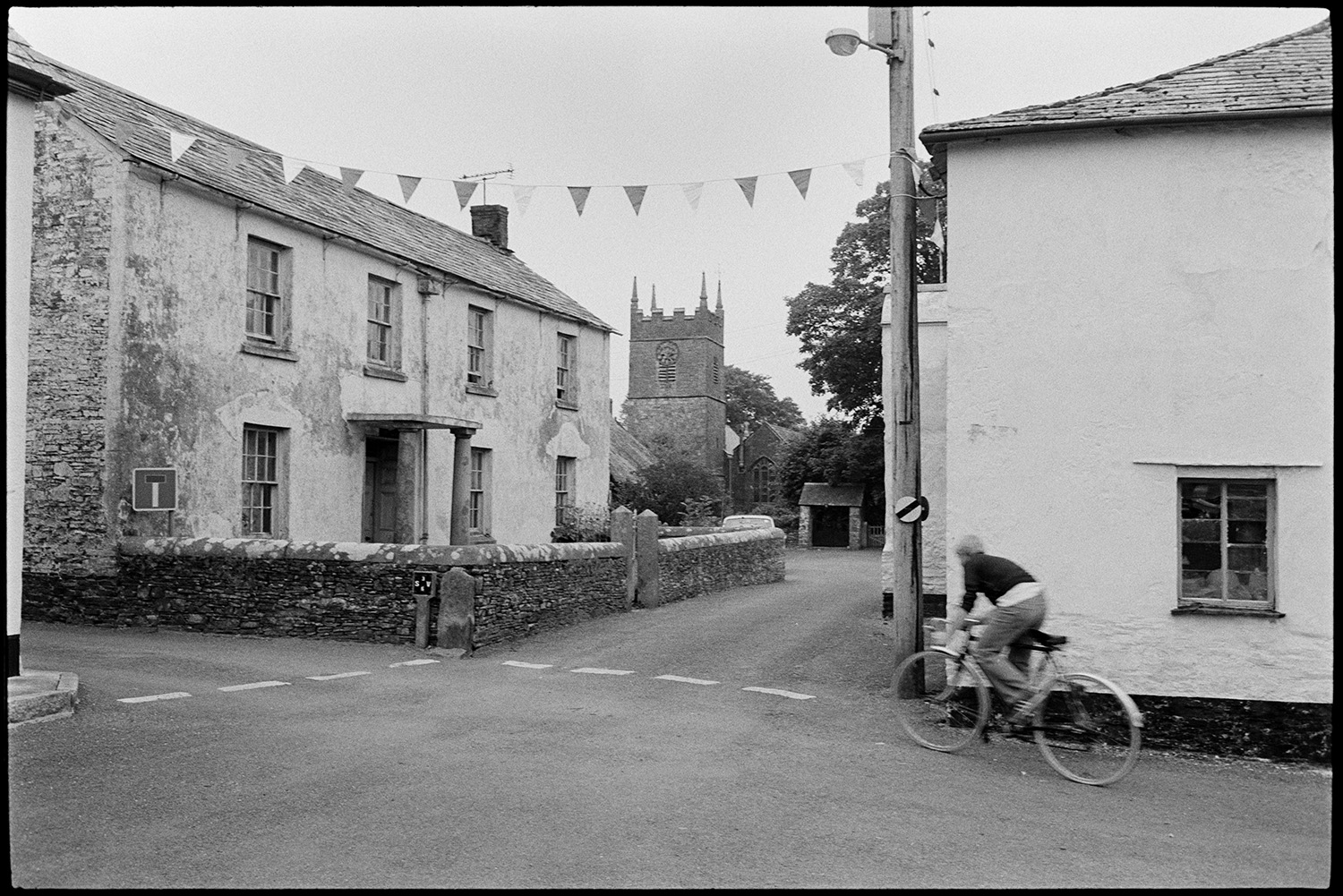 Street scenes, cars parked and bicycles, house with porch with stone pillars. 
[A person cycling past a road junction and houses in Northlew. The street is decorated with bunting. One of the houses has a porch supported by stone pillars. Northlew Church tower can be seen in the background.