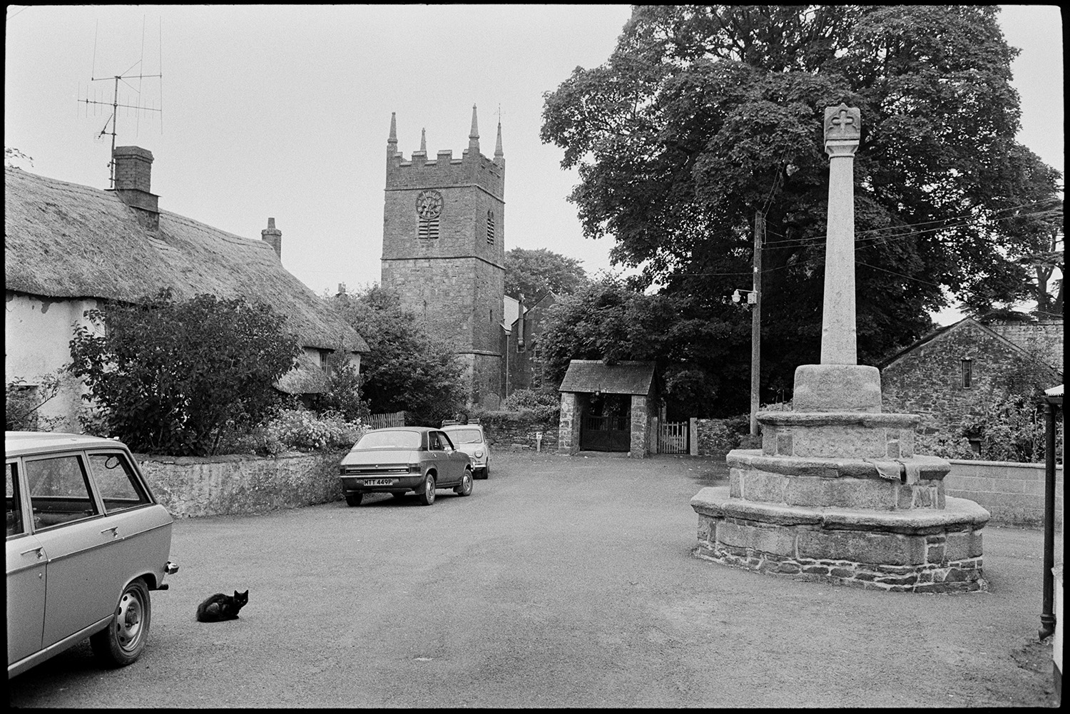 Street scenes, cars parked and bicycles, house with porch with stone pillars. 
[A street outside Northlew Church with a war memorial. A thatched cottage and parked cars are on the side of the street. A cat is sat by one of the cars. The gate to the church can also be seen.]