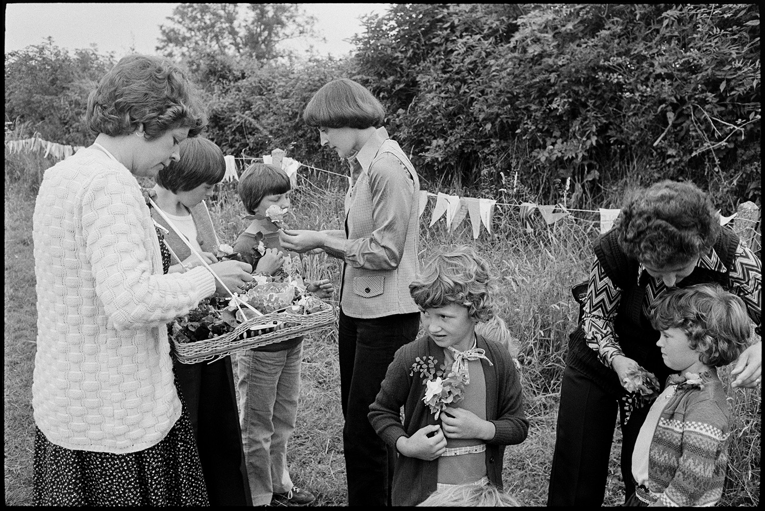 Village sports day, speeches, eating tea in hall, races, people chatting. 
[Women and children buying small floral bouquets or button holes from a woman at Roborough Sports Day. Bunting is hung along the hedge in the background.]