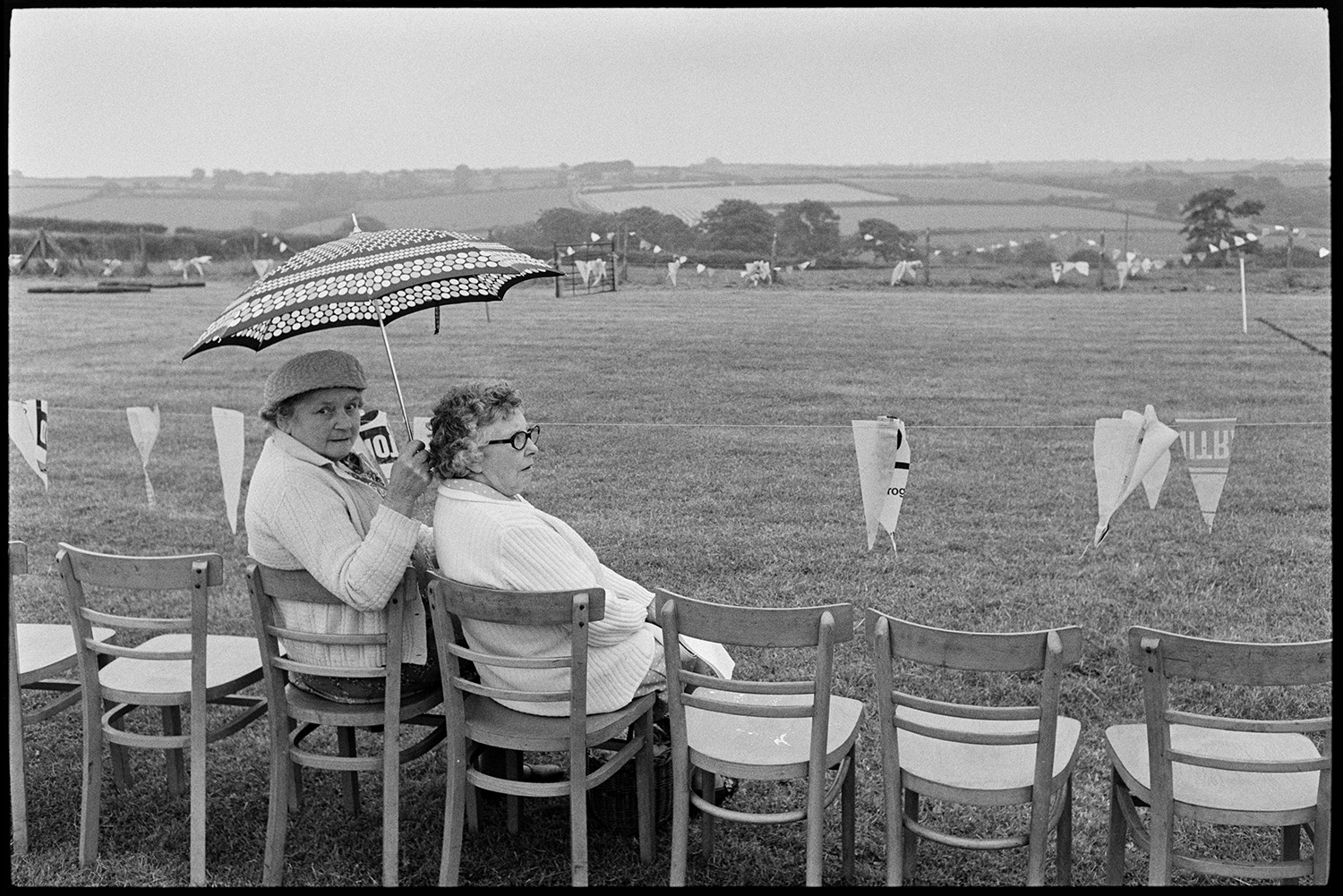 Village sports day, speeches, eating tea in hall, races, people chatting. 
[Two women sat on chairs waiting to watch races at Roborough Sports Day. One of the women is holding an umbrella. The field is decorated with bunting.]